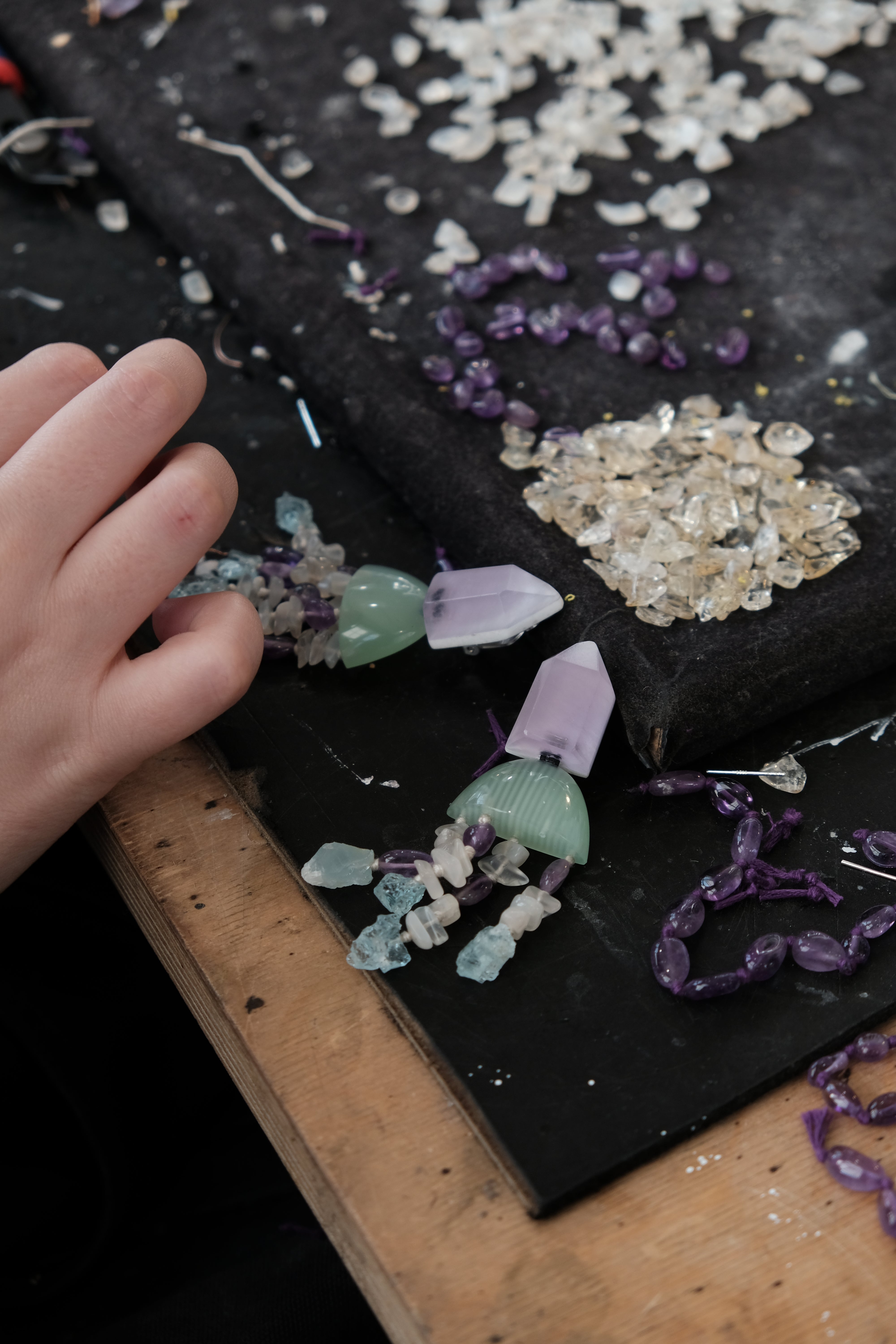 A close-up of a person's hand arranging handmade beaded jewelry made of translucent crystals and purple beads on a black mat over a wooden table.