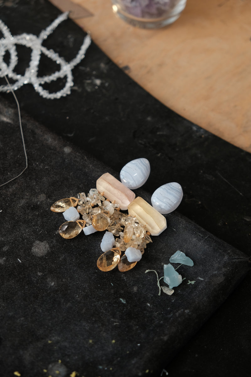A handmade beaded and crystal jewelry piece displayed on a black fabric surface, featuring white marbled beads, pale pink and cream rectangular beads, clear faceted crystals, and amber-colored teardrop beads.
