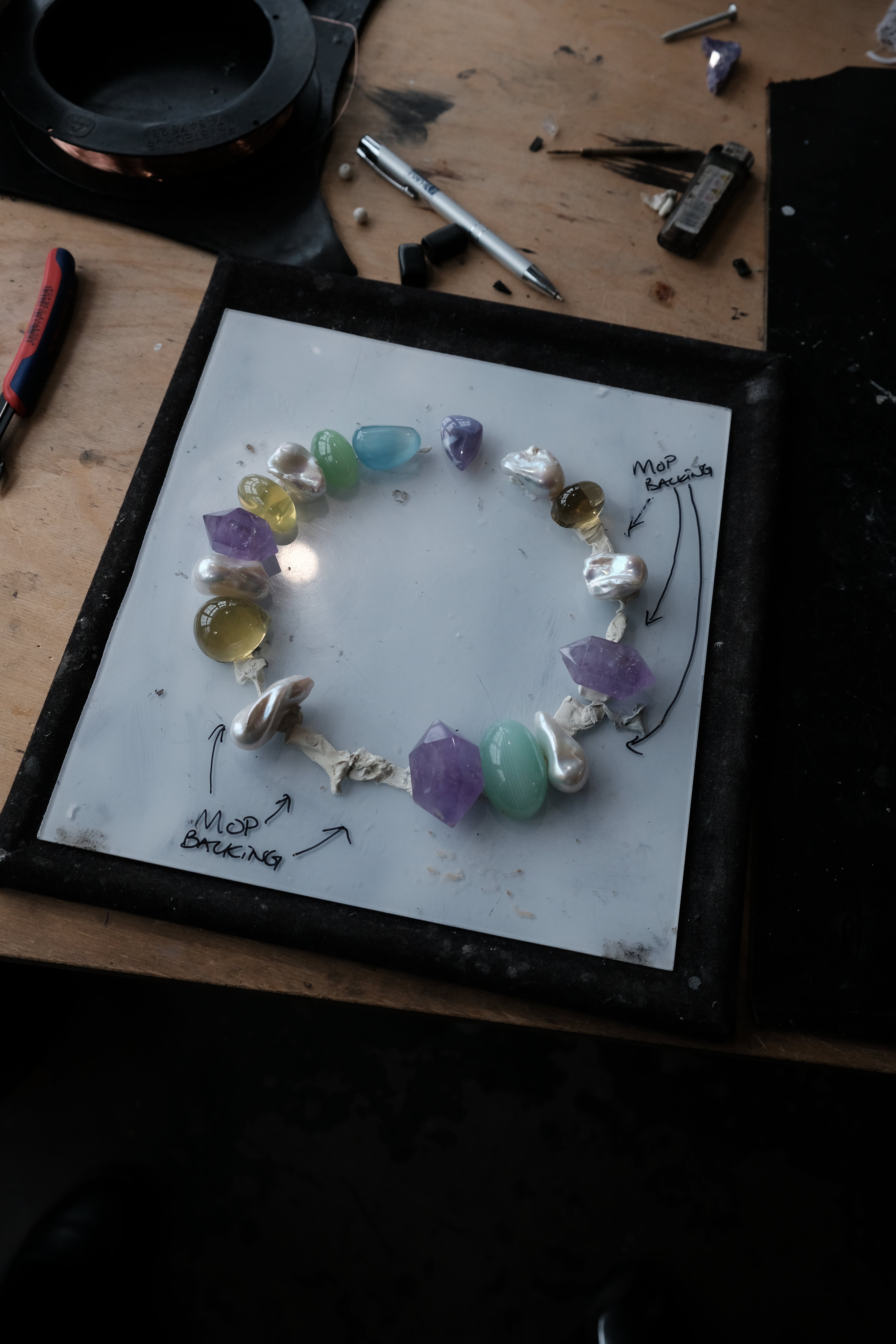 A handmade beaded necklace made of colorful polished gemstones and white irregular pearls, displayed on a white square board with handwritten notes, set on a wooden workbench surrounded by jewelry-making tools.
