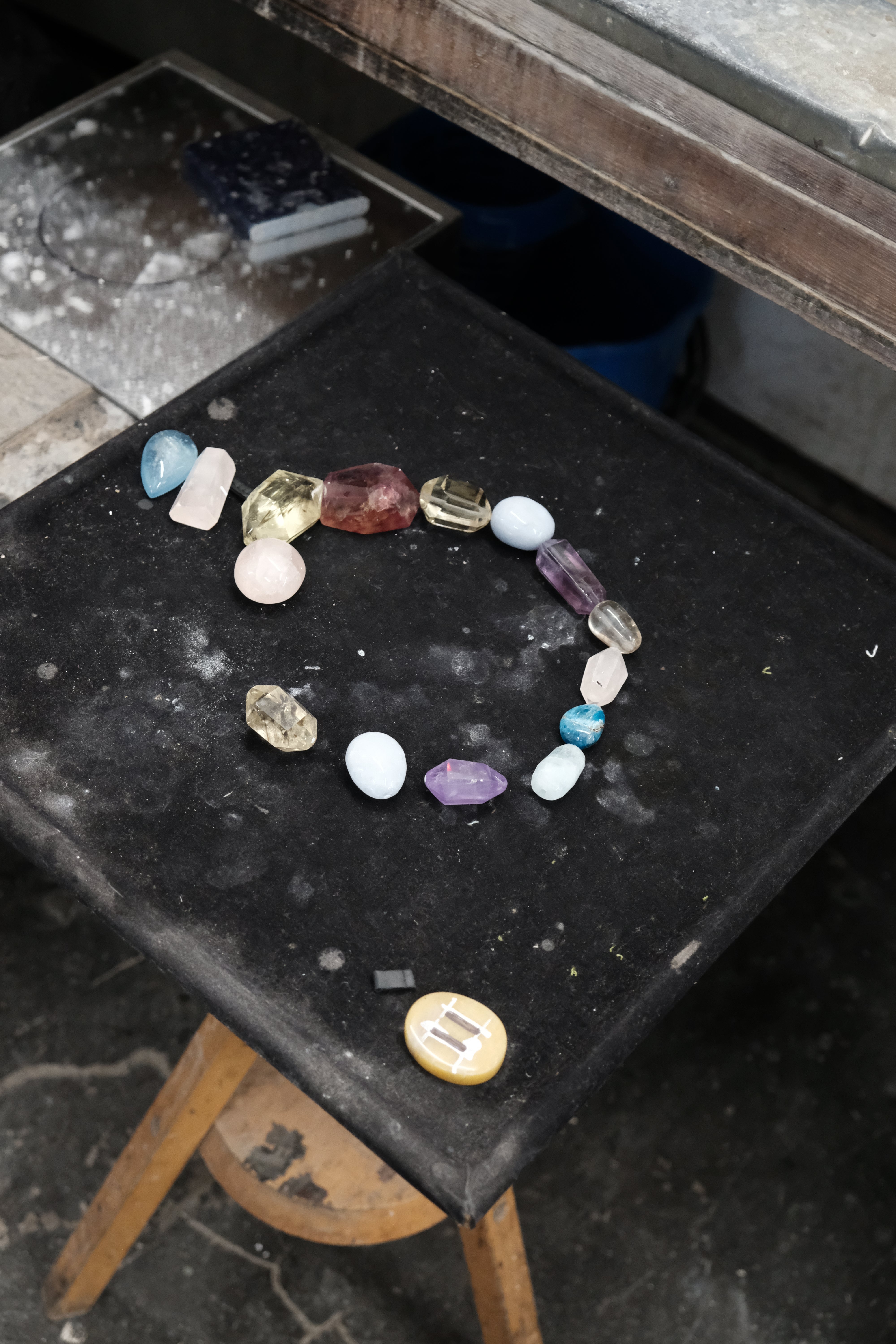 A multicolor beaded necklace featuring faceted gemstone beads in shades of pink, light blue, purple, amber, and clear, with a teardrop-shaped light blue stone pendant hanging from the center, laying on a black workbench.