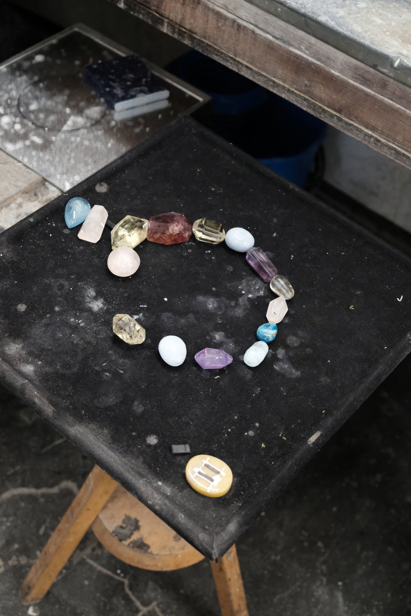 A multicolor beaded necklace featuring faceted gemstone beads in shades of pink, light blue, purple, amber, and clear, with a teardrop-shaped light blue stone pendant hanging from the center, laying on a black workbench.
