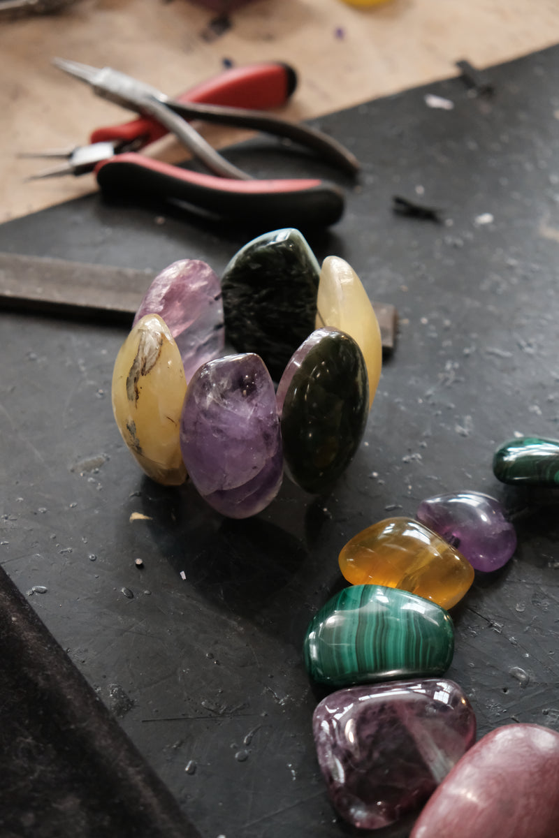A cluster of polished tumbled gemstones in purple, yellow, green, and black arranged on a dark workbench, with pliers and other tools blurred in the background.