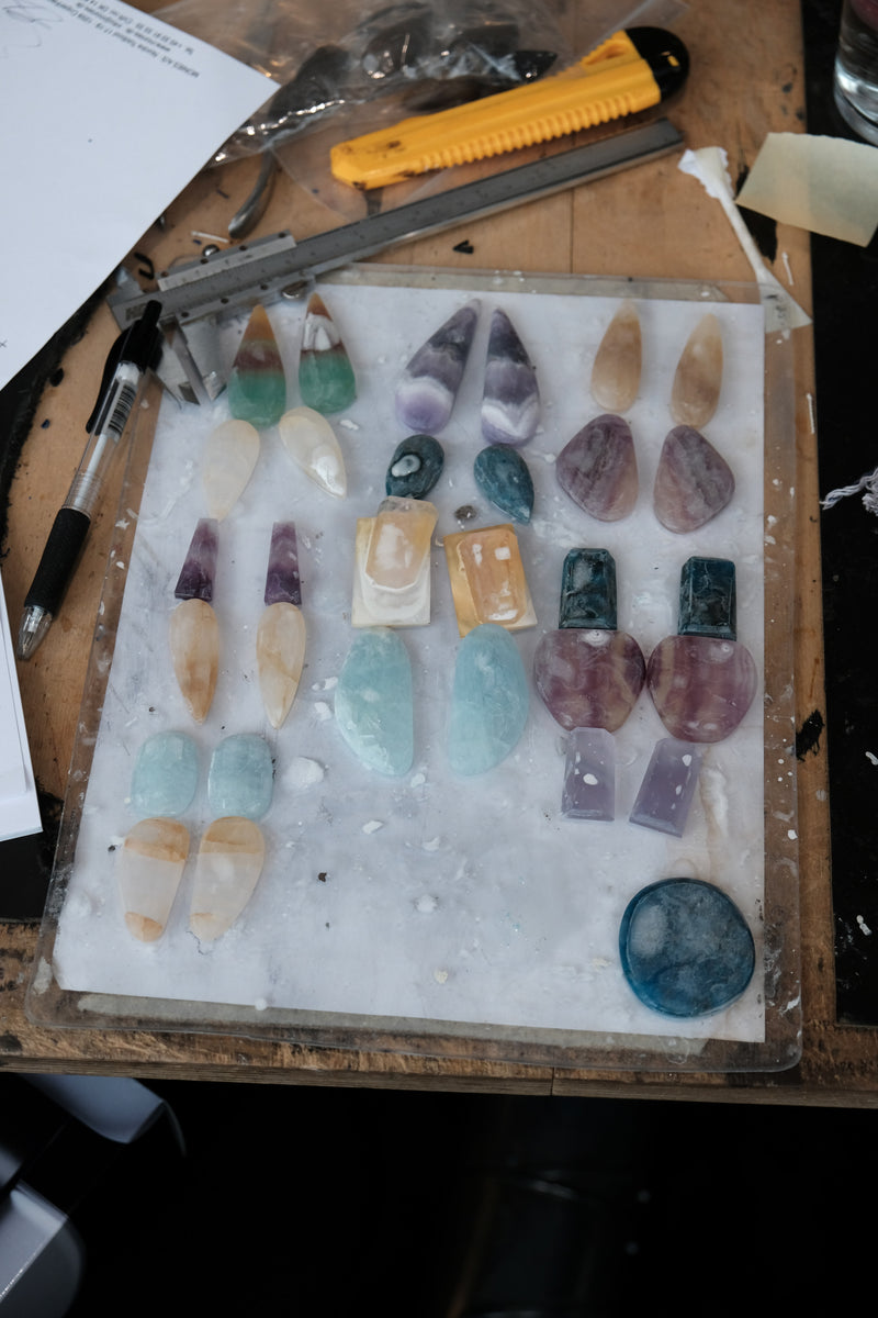 A collection of polished gemstones and crystal chips arranged on a white sheet over a wooden workbench, with tools like calipers and a yellow utility knife visible around the edges.