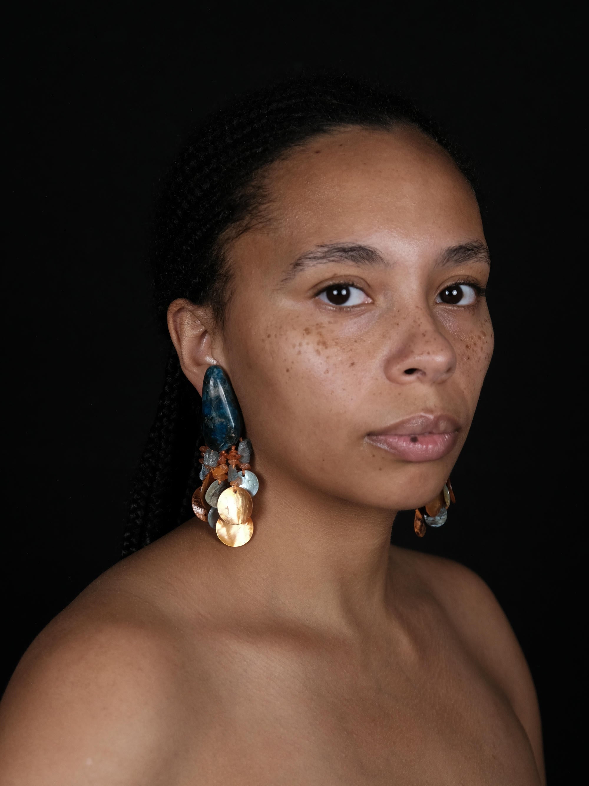 A close-up portrait of a woman with braided hair wearing large, colorful statement earrings against a solid black background.