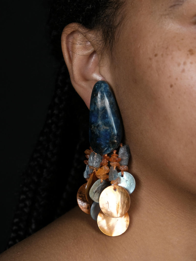 A close-up of a person's ear wearing a large statement dangle earring made from a polished dark blue stone and multiple dangling metallic and translucent discs in blue, copper, and silver tones.