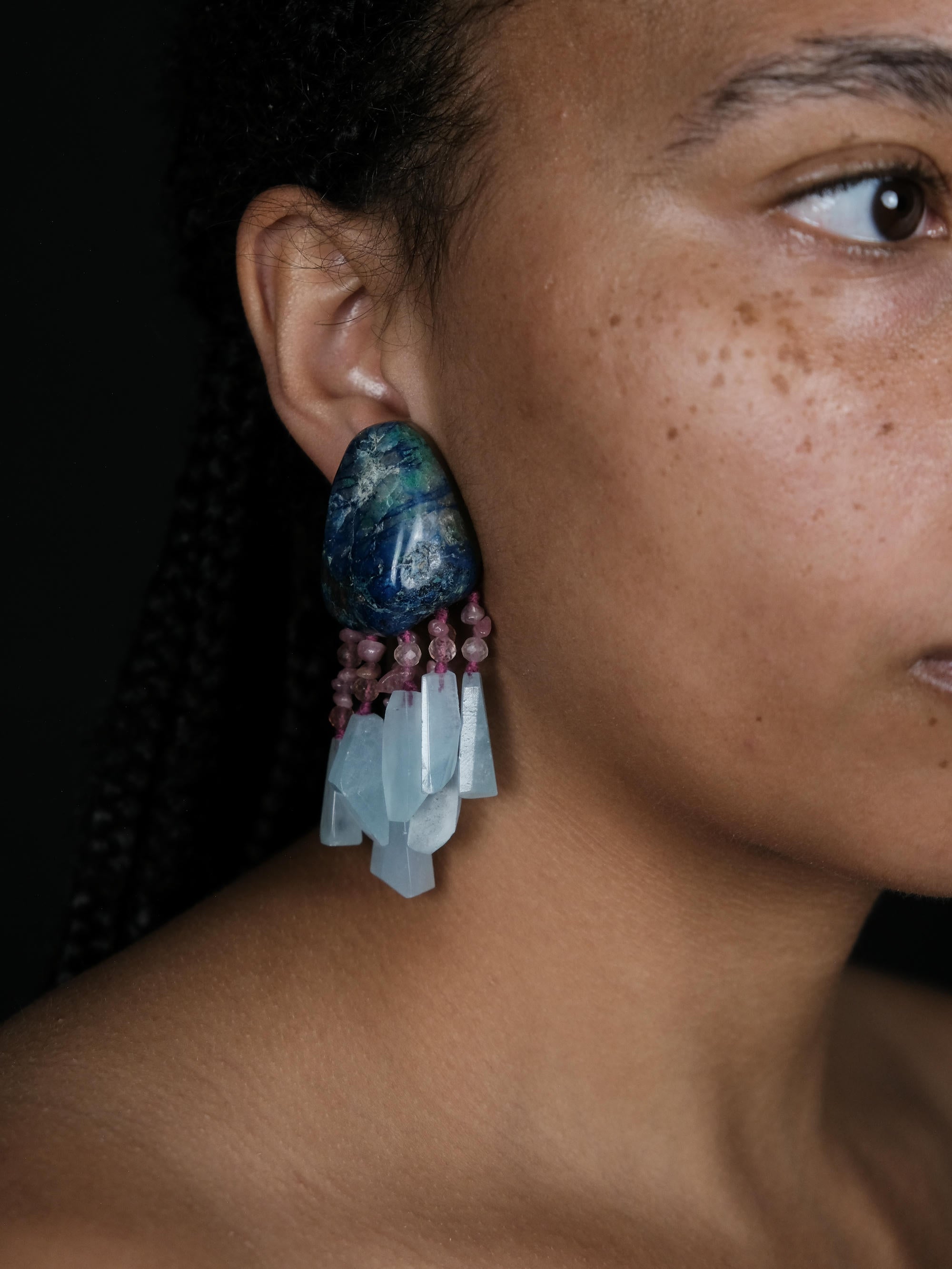 A close-up of a person's ear wearing a large statement earring. The earring features a polished blue and green marbled top stone, small dusty pink beads, and a cluster of translucent pale blue crystal points.