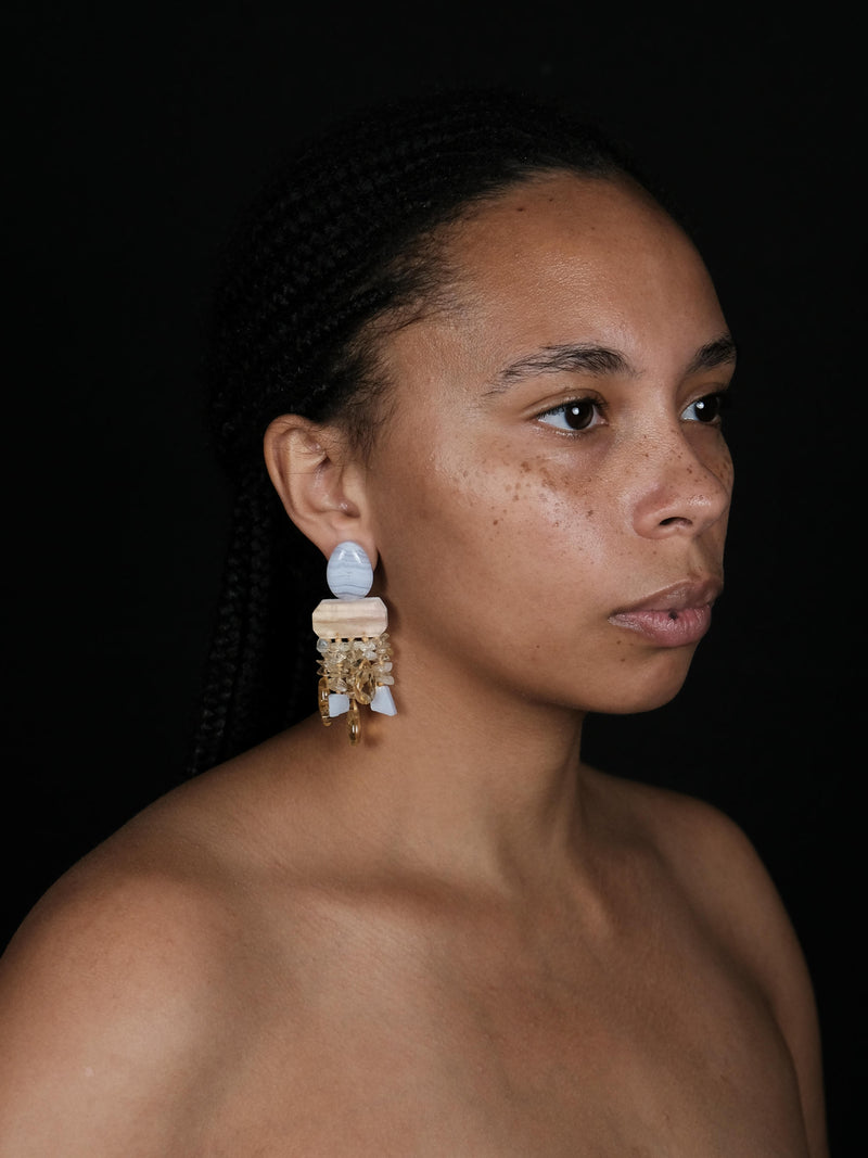 A woman wearing dangle earrings in purple and yellow stones. Displayed on a white background