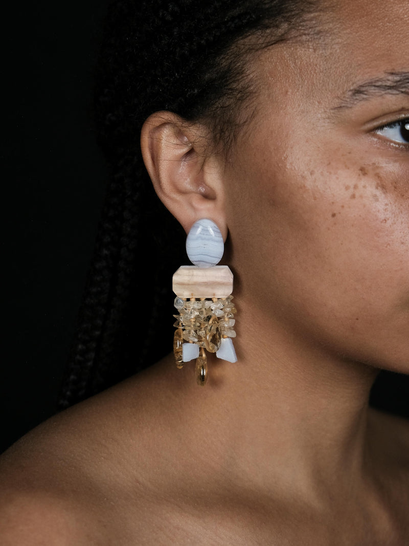 A woman wearing dangle earrings in purple and yellow stones. Displayed on a white background
