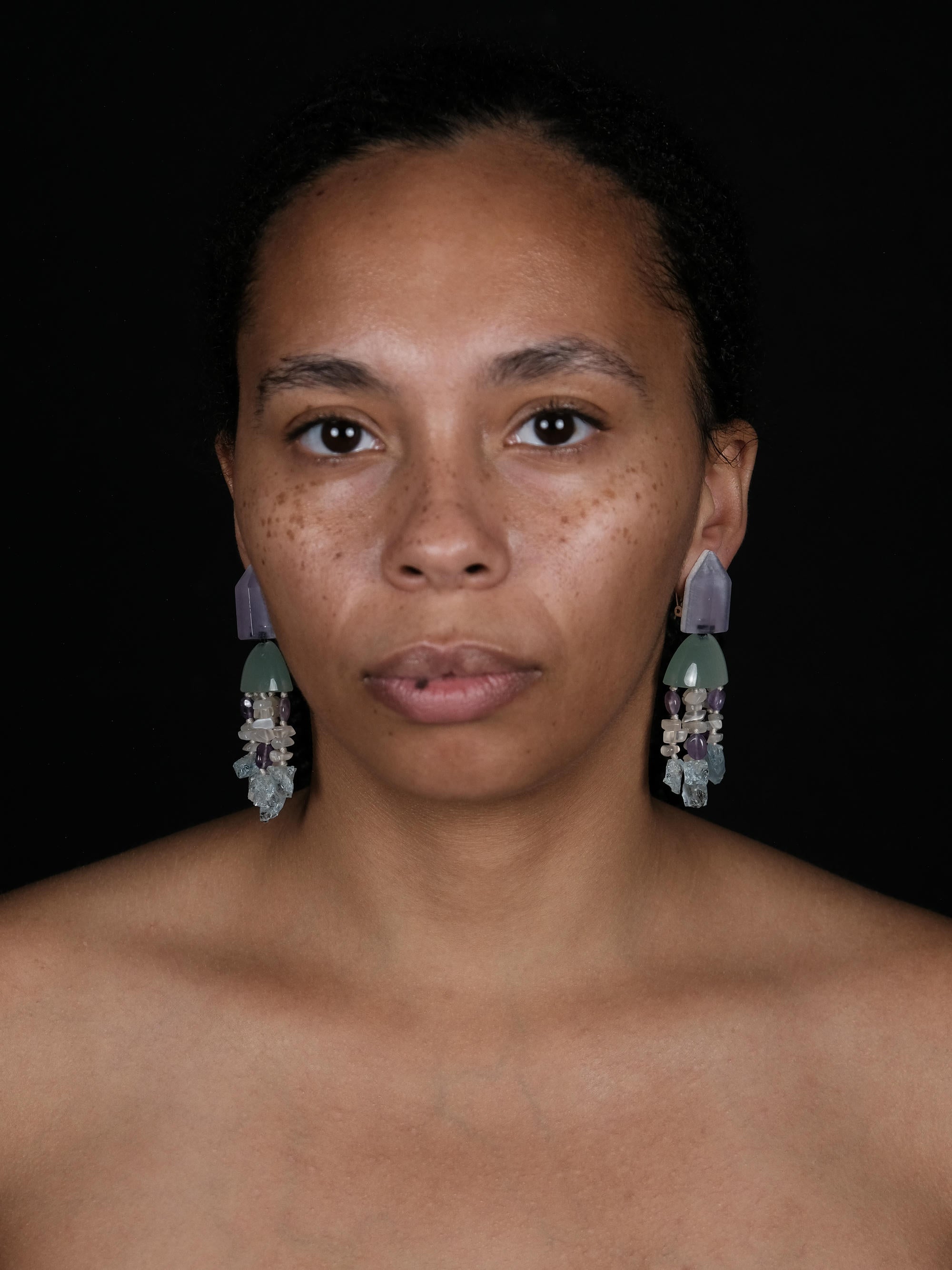 A woman wearing dangle earrings with a purple and green top and beige, purple and blue tassles underneath. Displayed on a white background. 