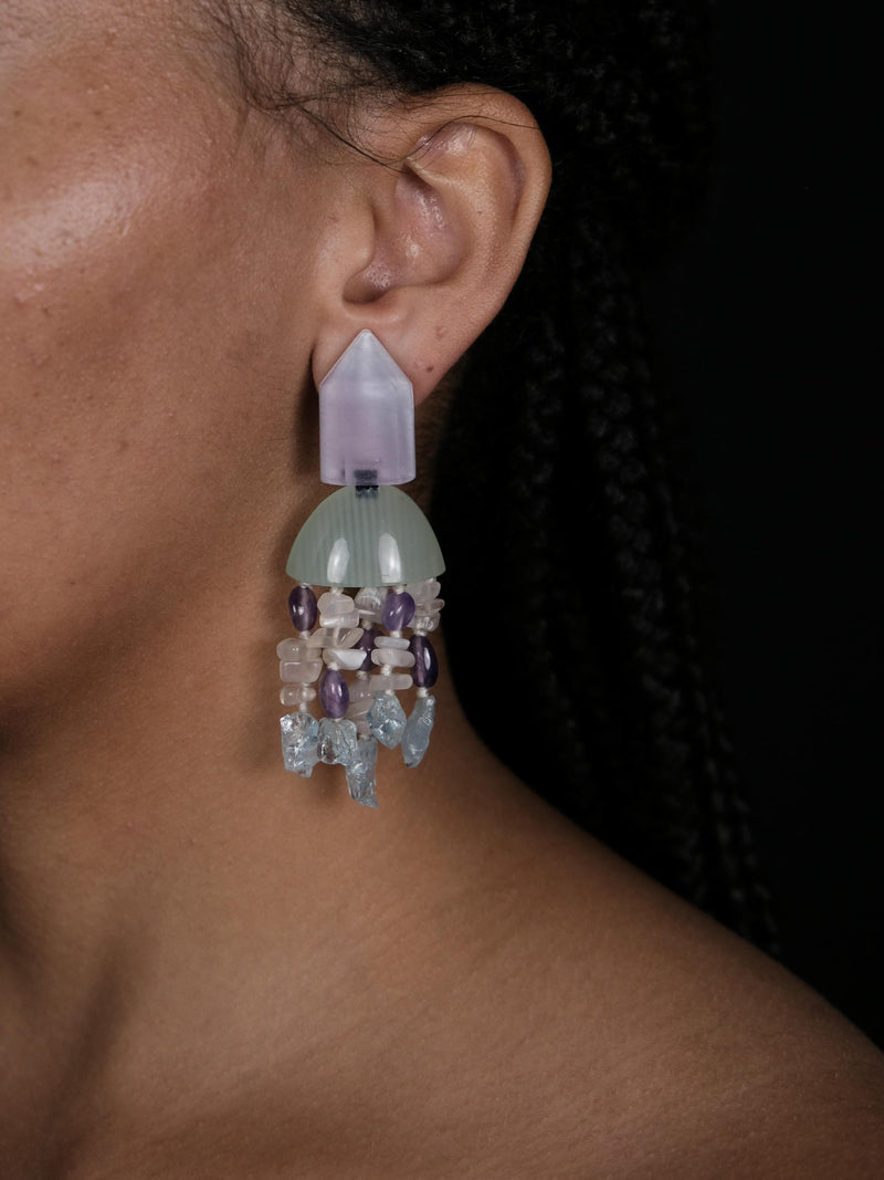 A woman wearing dangle earrings with a purple and green top and beige, purple and blue tassles underneath. Displayed on a white background. 