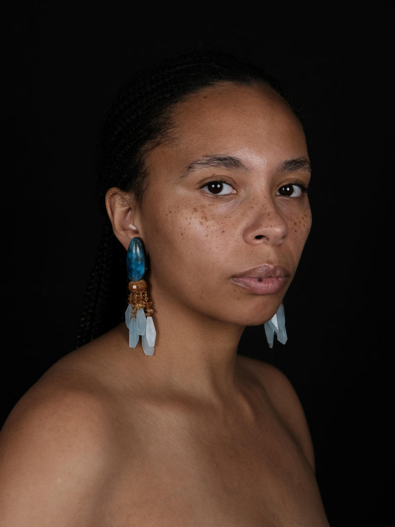 A woman wearing A pair of statement dangle earrings featuring polished blue-green teardrop beads, light blue crystal clusters, and textured orange-brown metallic accents.