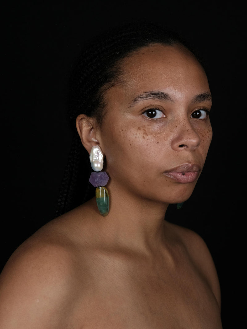 A woman wearing Baroque pearl dangle earrings with purple and yellowish green stones underneath. Displayed on a white background