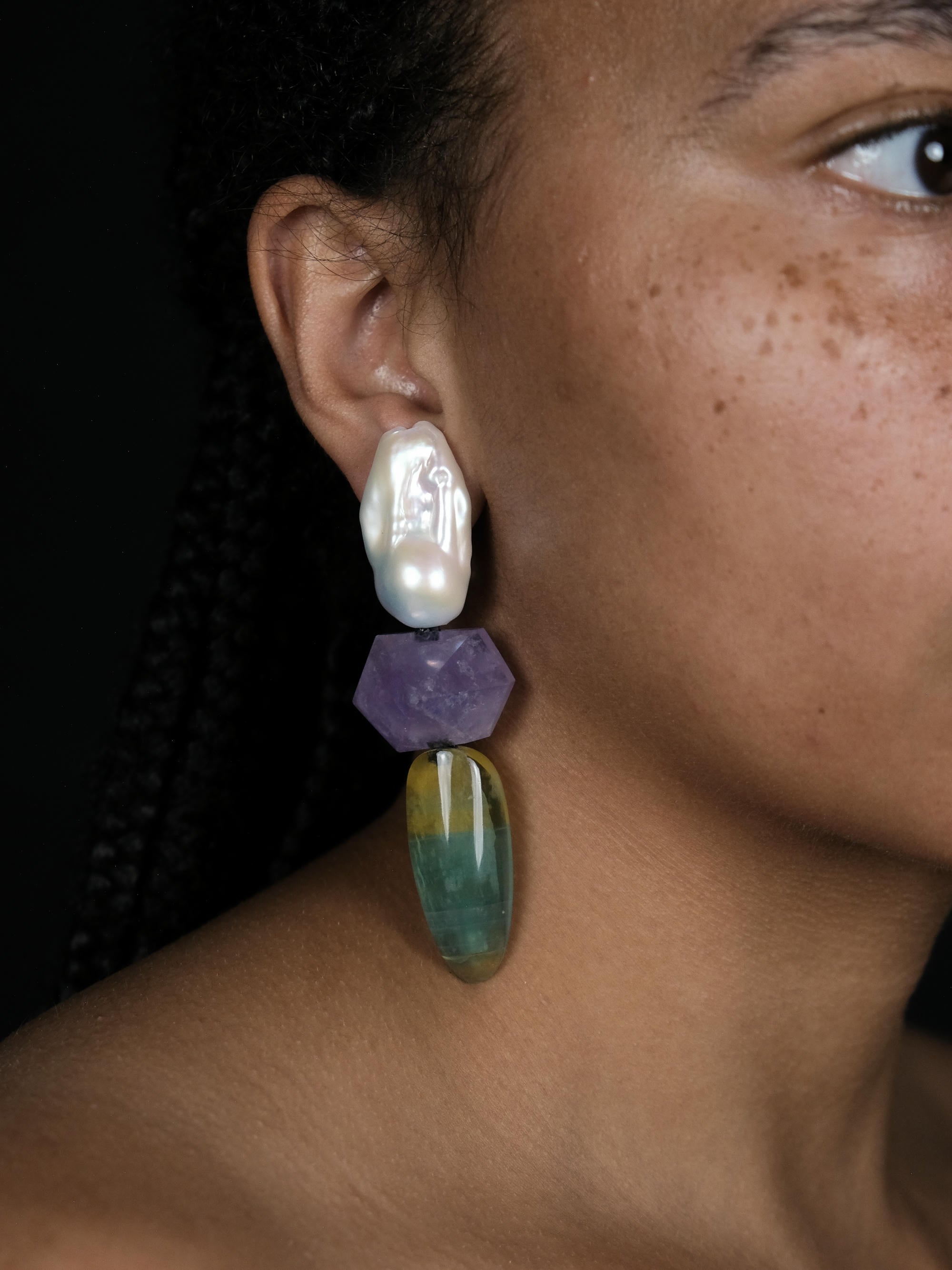 A close up of a woman wearing Baroque pearl dangle earrings with purple and yellowish green stones underneath. Displayed on a white background