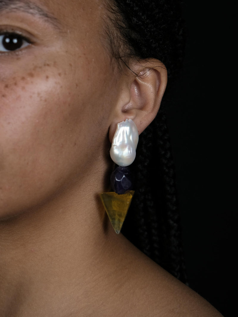 A close up of a woman wearing baroque pearl earrings with a faceted purple and yellow stones underneath. Displayed on a white background