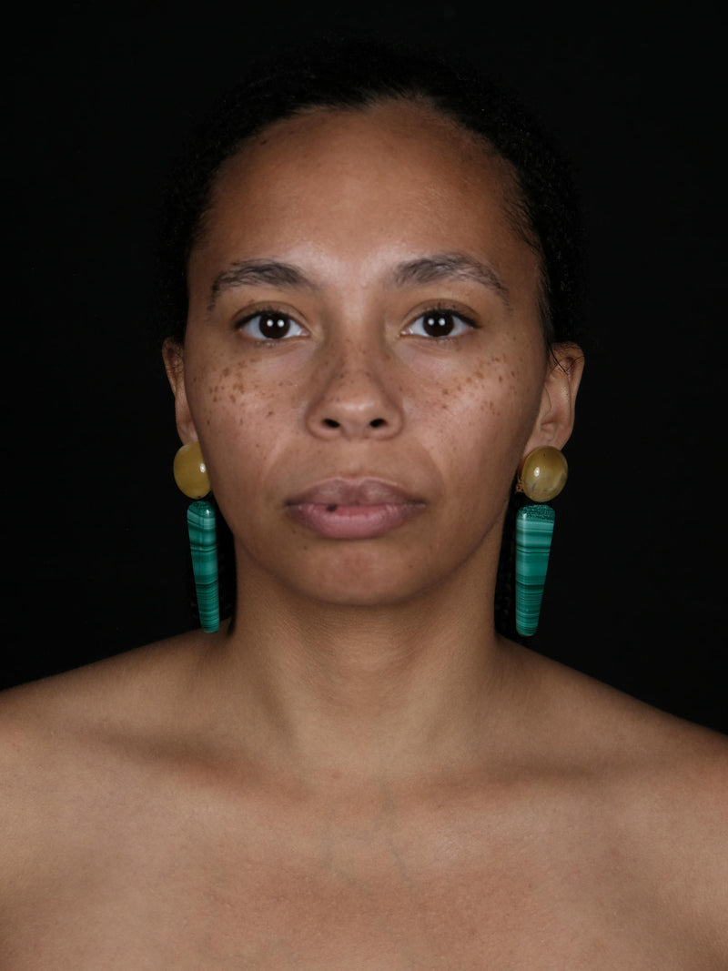 A close-up portrait of a woman wearing long, chunky turquoise and yellow drop earrings against a solid black background.