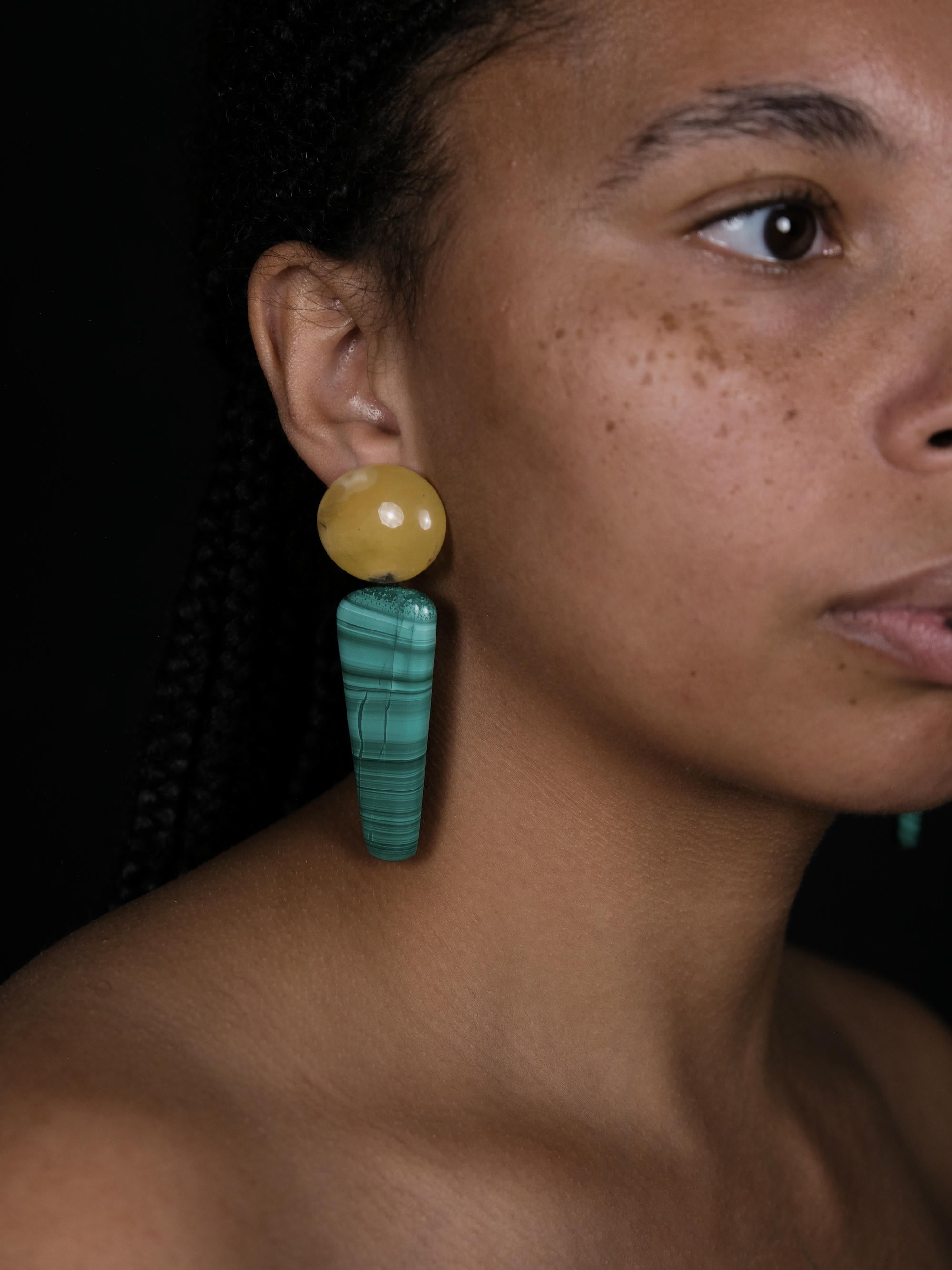A close-up of a model wearing a long dangle earring with a round yellow top and a turquoise-green striped drop, set against a dark background.