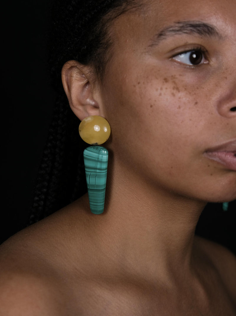 A close-up of a model wearing a long dangle earring with a round yellow top and a turquoise-green striped drop, set against a dark background.