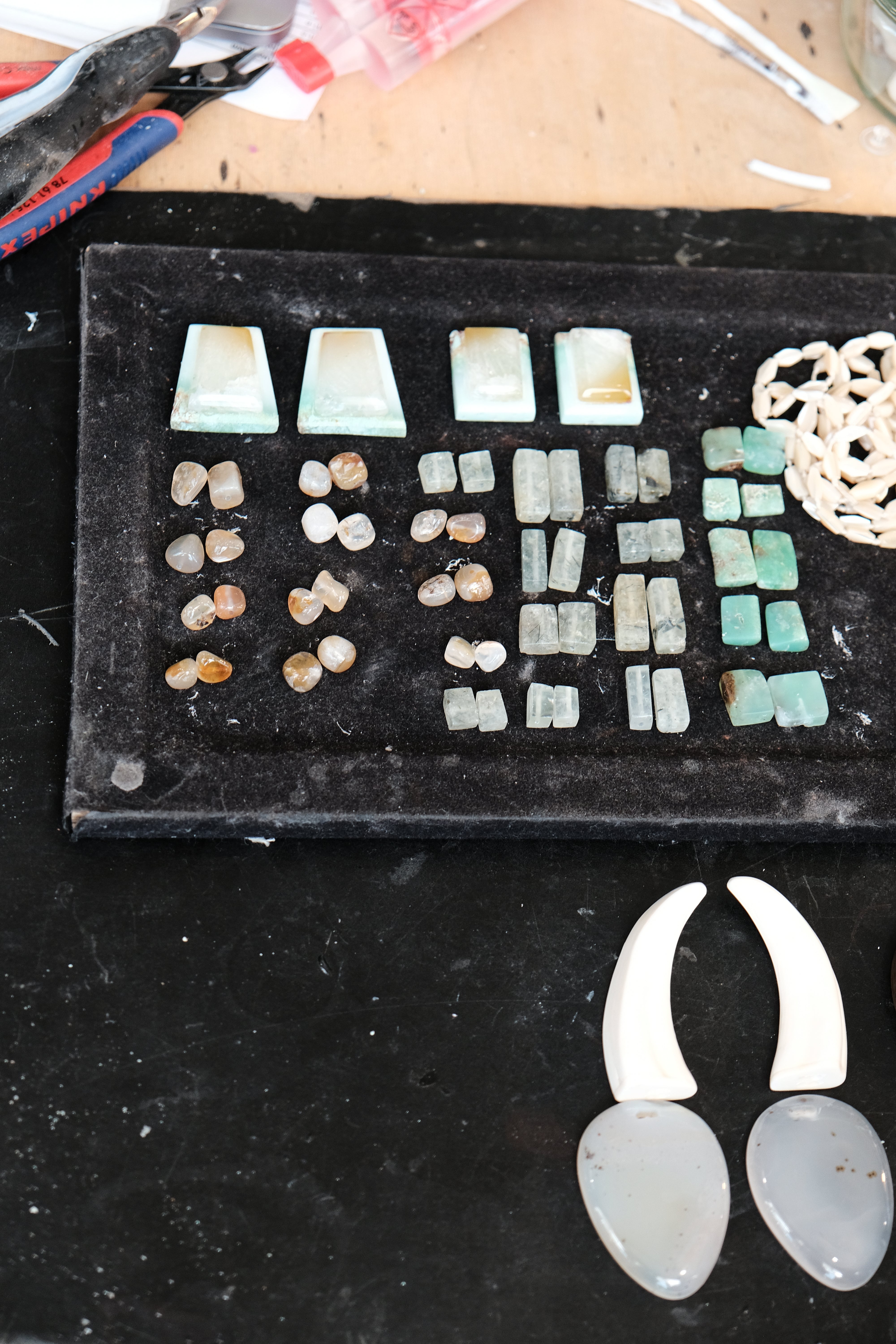 An assortment of raw gemstone beads and pendants arranged on a black display tray, with two larger off-white teardrop pendants in the foreground on a dark surface.