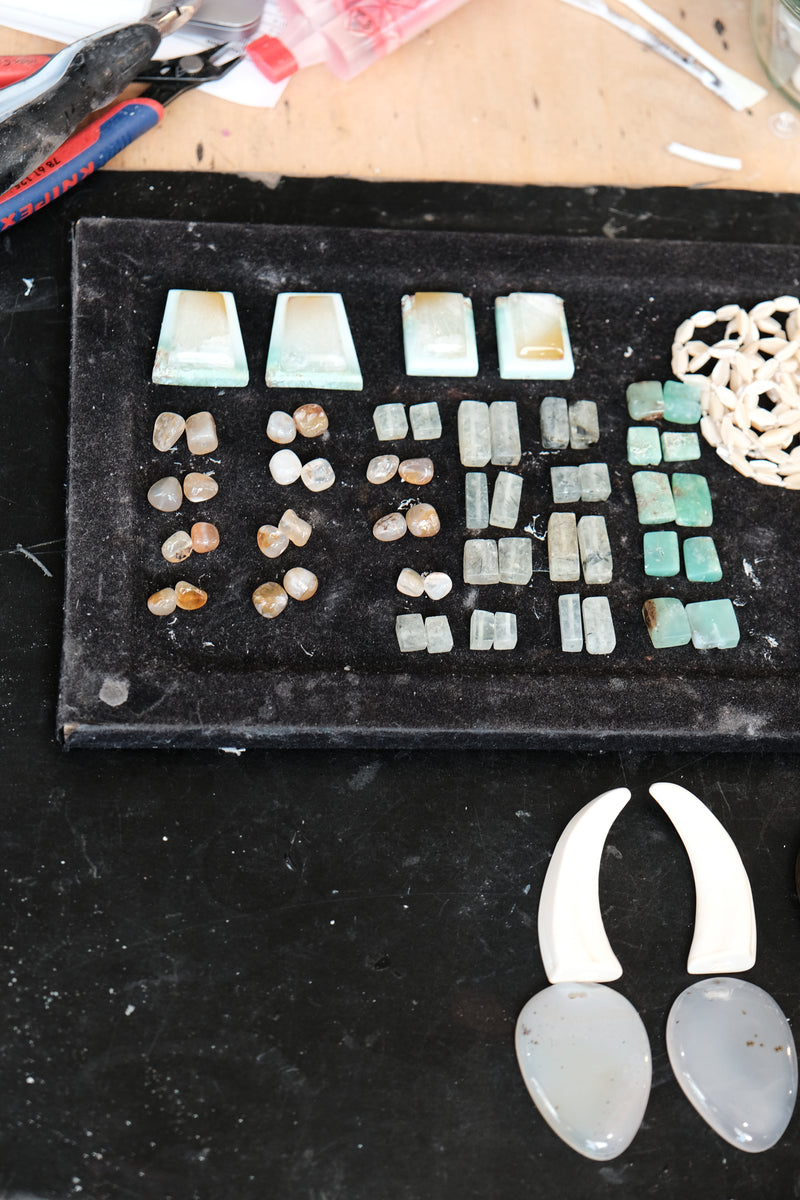 An assortment of raw gemstone beads and pendants arranged on a black display tray, with two larger off-white teardrop pendants in the foreground on a dark surface.