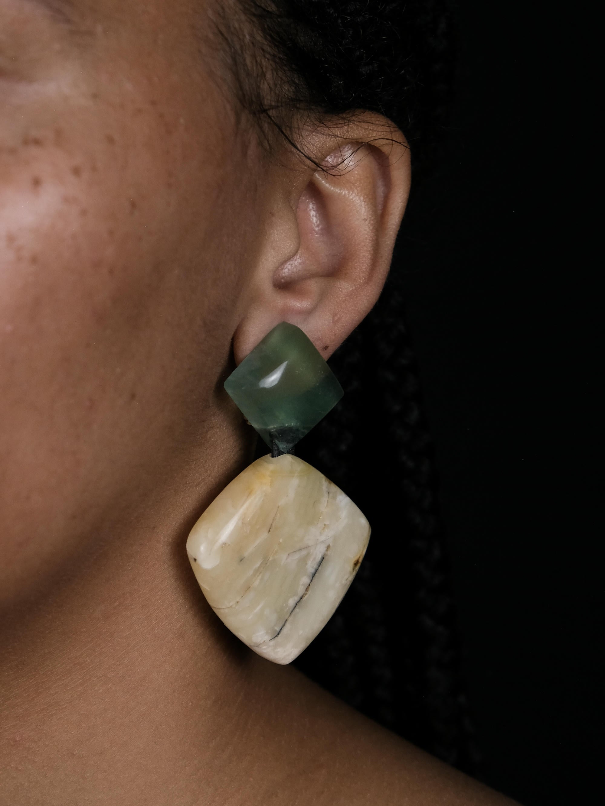 A close-up of a person's ear wearing a large statement earring made of two stacked organic-shaped stones, one dark green and one cream-colored with natural marbling, set against a dark background.
