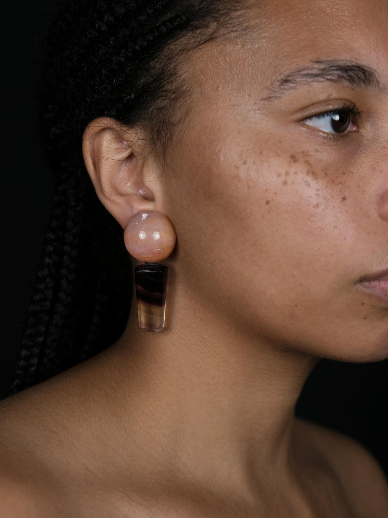 A close-up side profile of a woman wearing a unique statement earring. The earring features a large, round, pale pinkish-beige top bead and a dangling, tapered clear resin section with a dark brown middle band, set against a solid black background.