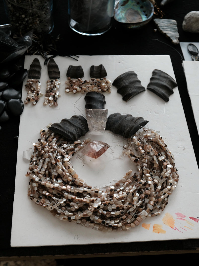 A handmade beaded necklace and matching drop earrings displayed on a white board. The jewelry features natural mother-of-pearl shell beads in shades of beige, cream, and brown, accented with clear crystals and a small pinkish crystal.