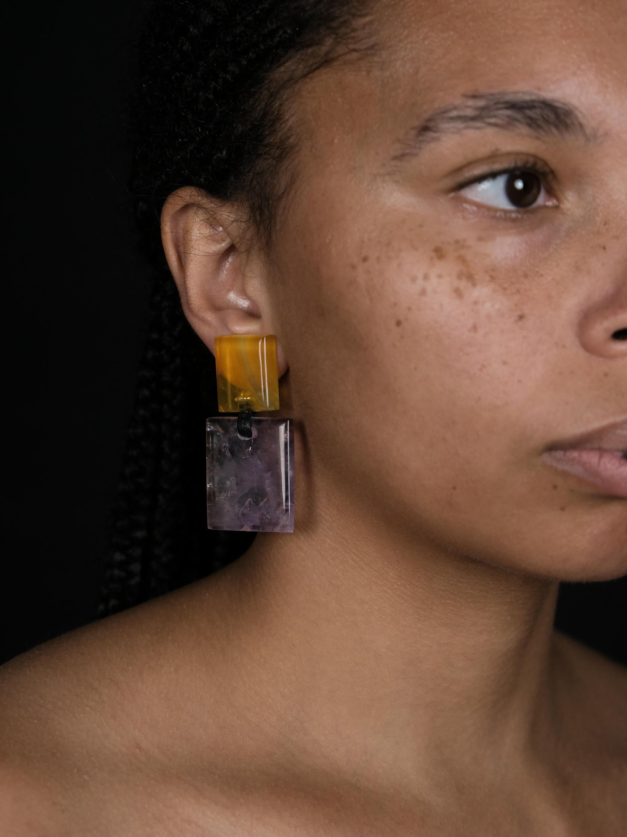 A woman wearing earrings made of a small yellow square and a larger purple square underneath, displayed on a white background.