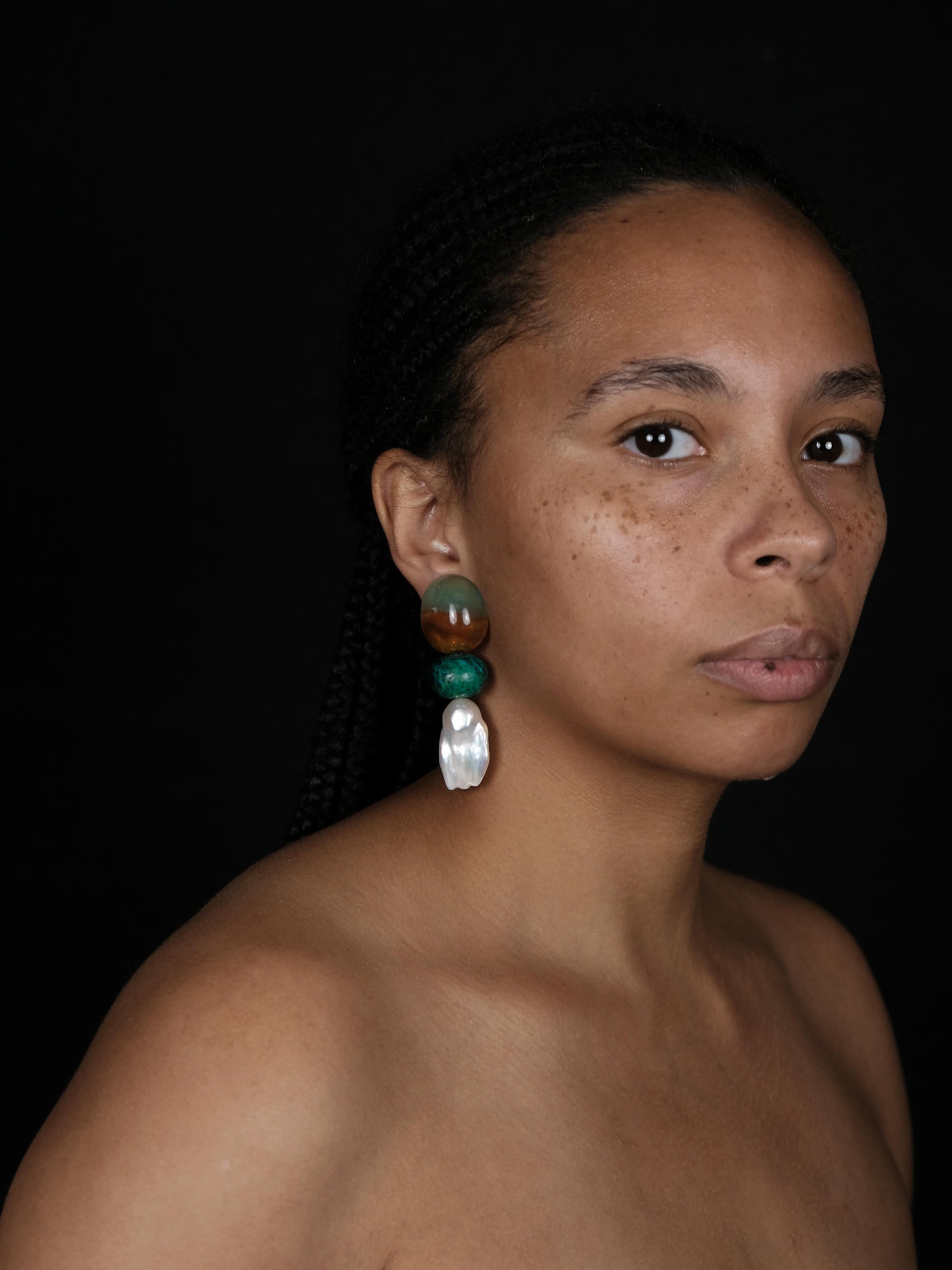 A woman wearing large statement dangle earrings in green, brown and baroque pearls.