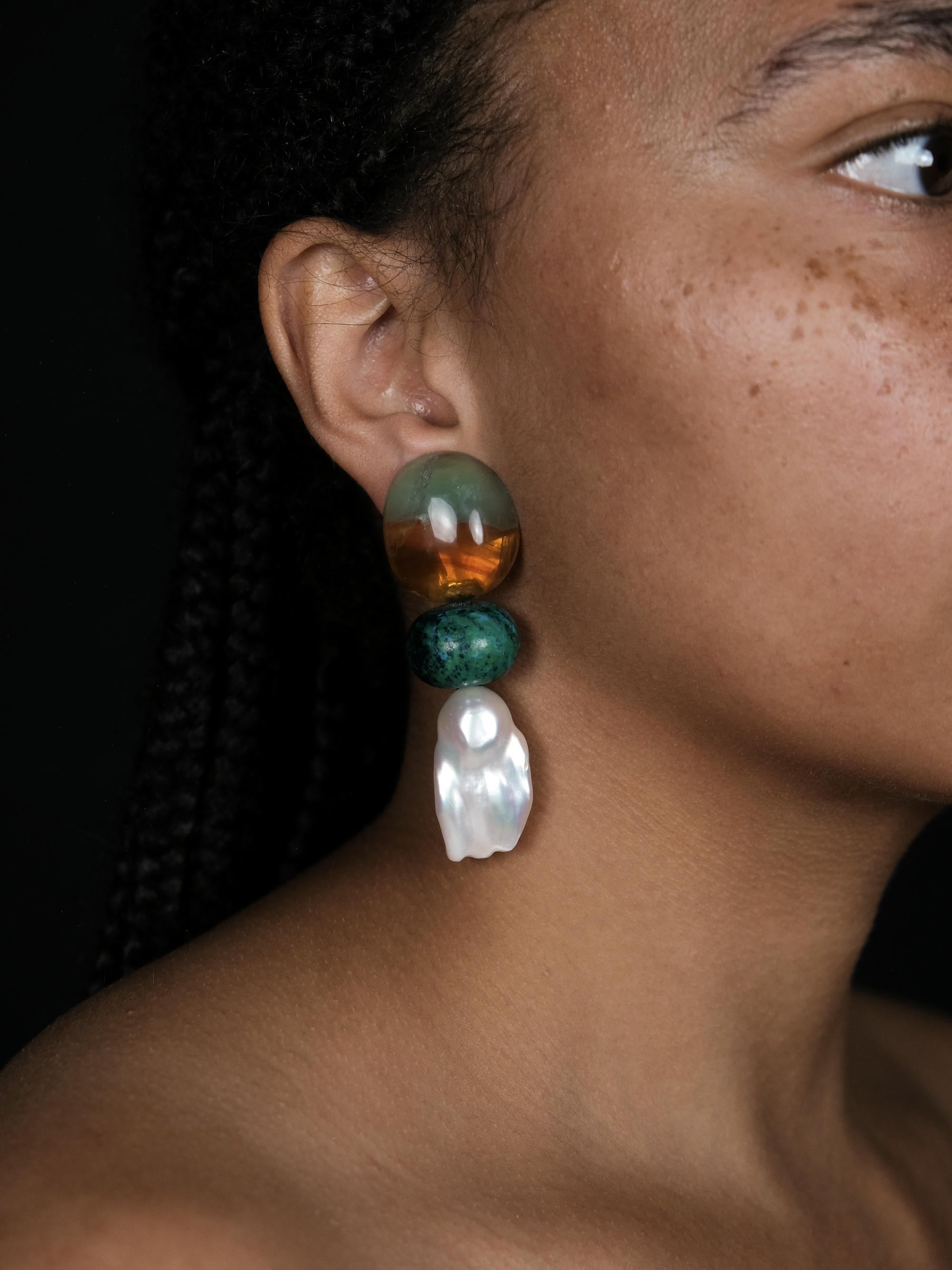 A close up of a woman wearing large statement dangle earrings in green, brown and baroque pearls.