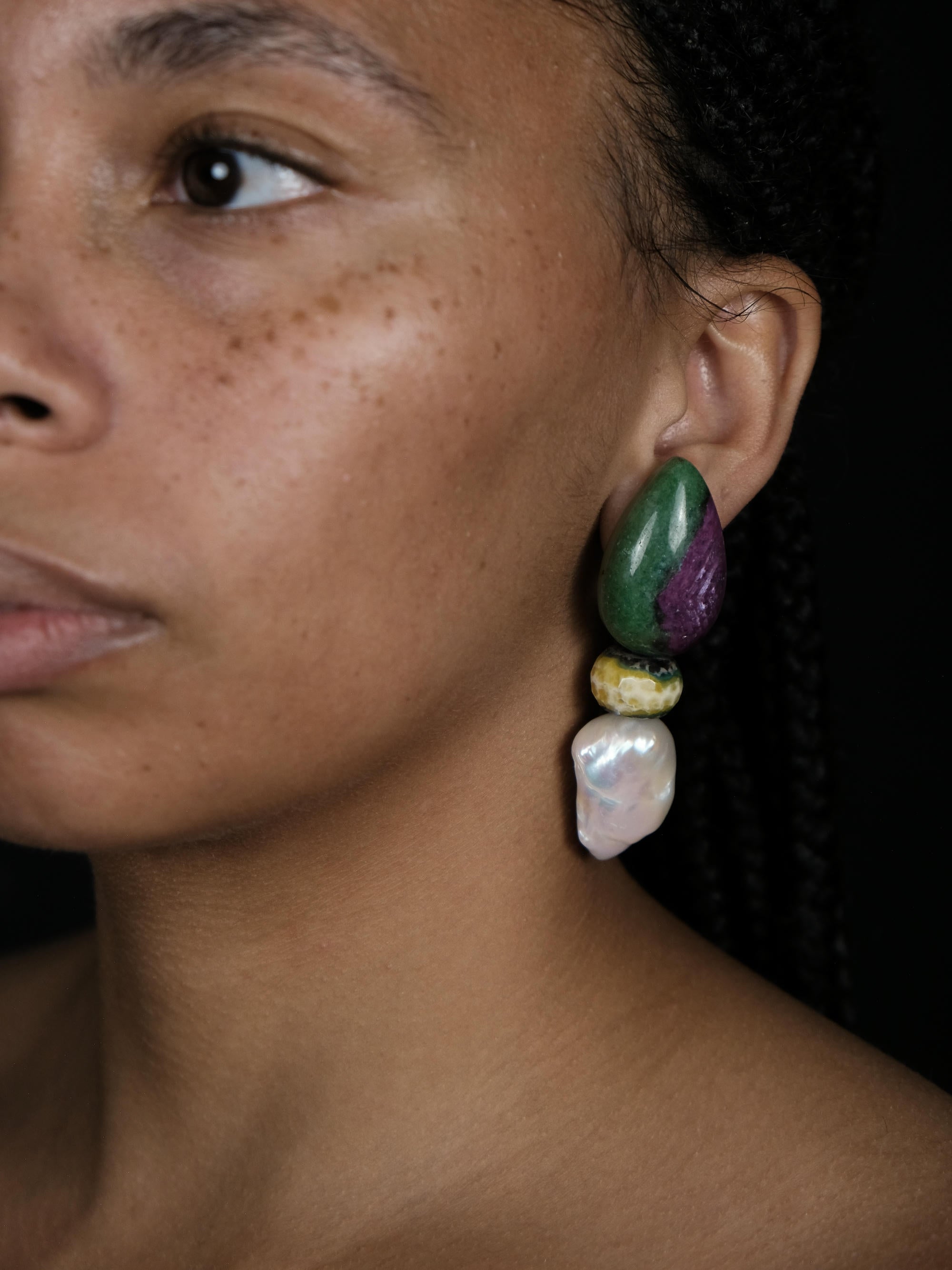 A close-up of a person's ear wearing a large, multi-colored statement earring made of polished green, purple, yellow, and white stones.