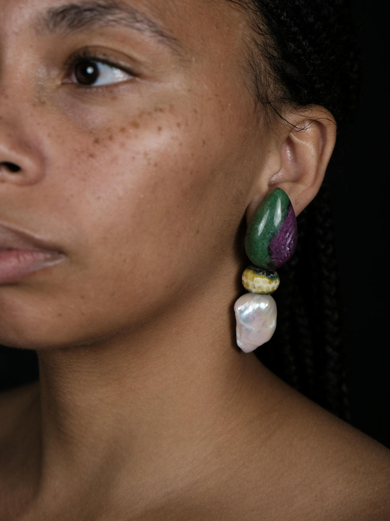 A close-up of a person's ear wearing a large, multi-colored statement earring made of polished green, purple, yellow, and white stones.