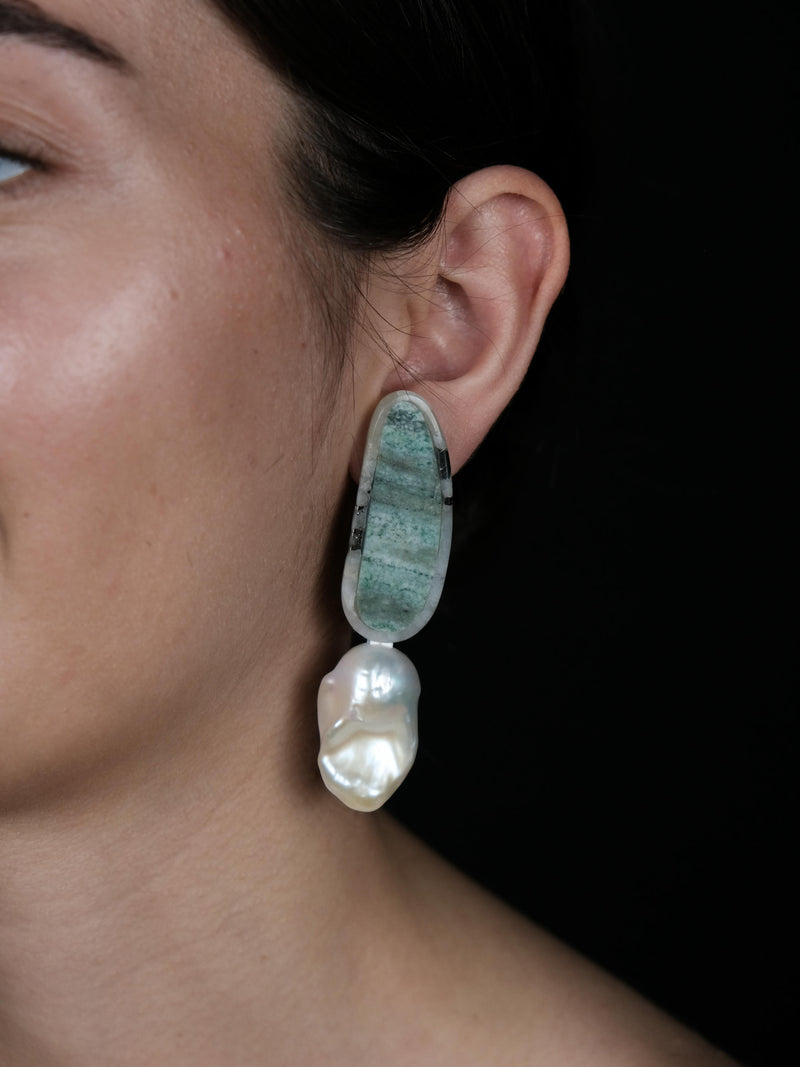 A close-up of a person's ear wearing a drop earring made of a green marbled stone on top of a large, irregular white baroque pearl, set against a solid black background.