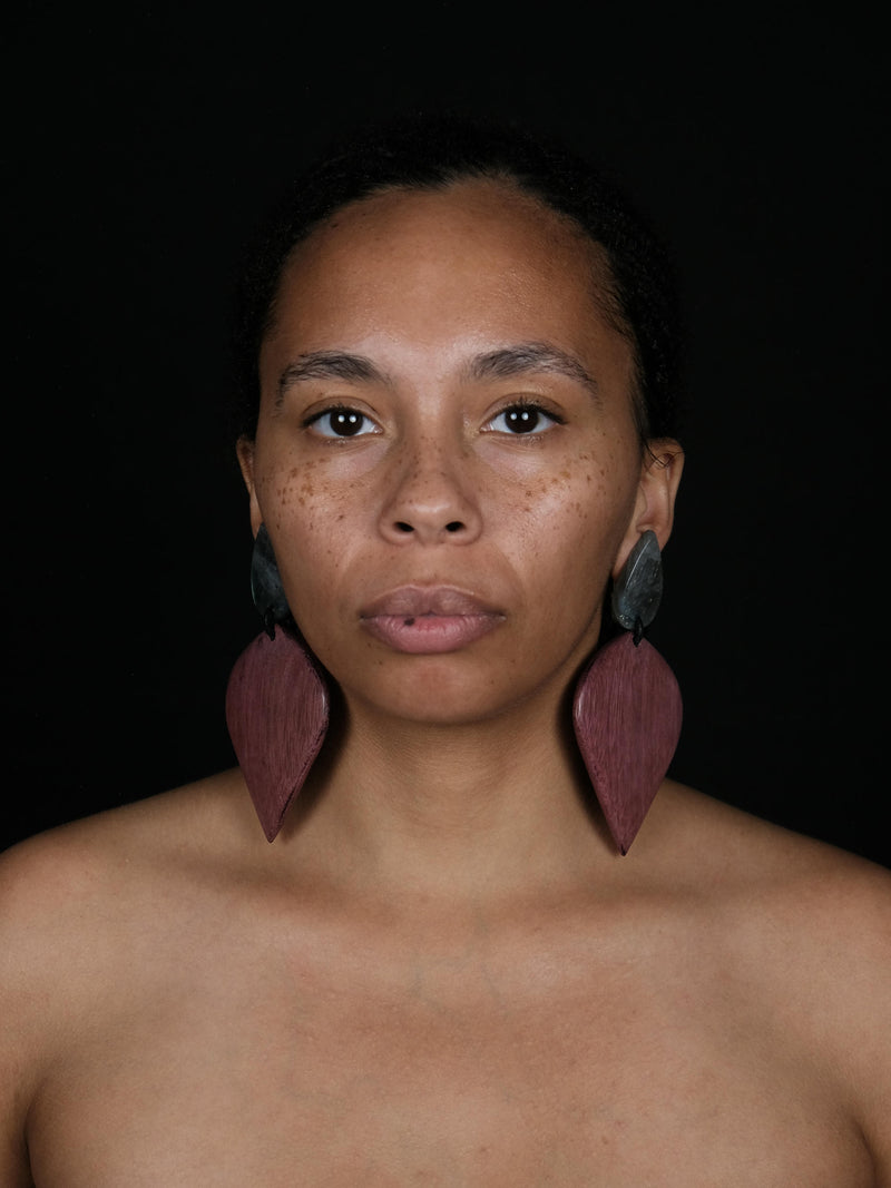A close-up portrait of a woman wearing large, teardrop-shaped wooden earrings in a dusty rose-brown color, set against a solid black background.