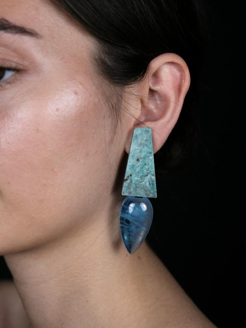 A close-up of a model's ear wearing a statement dangle earring. The earring features a textured, rectangular turquoise-blue stone top section and a polished, teardrop-shaped dark blue stone bottom section, set against a solid black background.