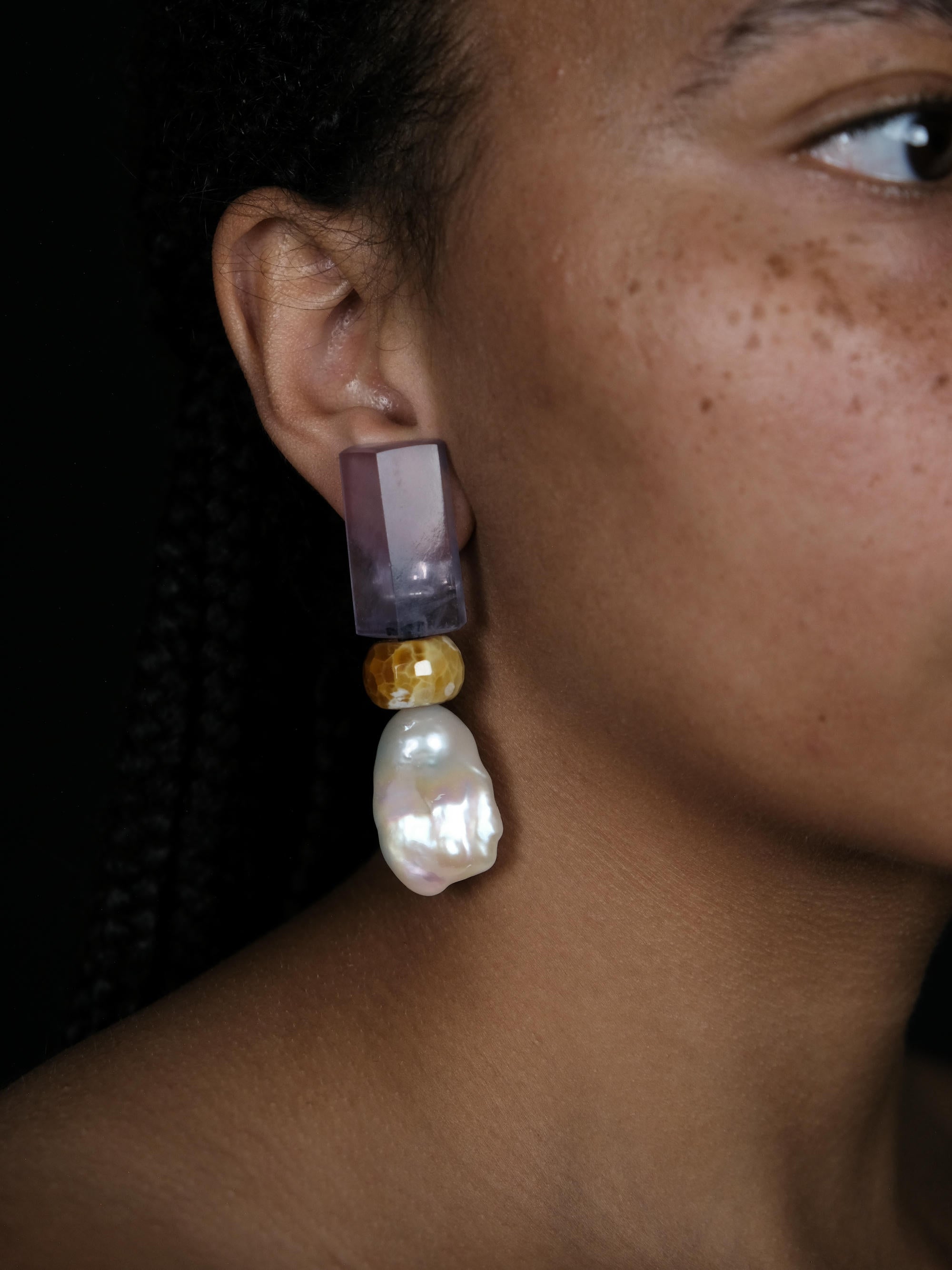 A close-up of a model's ear wearing a statement dangle earring. The earring features a translucent lavender-purple rectangular top, a small faceted gold bead, and a large, irregular white baroque pearl drop.