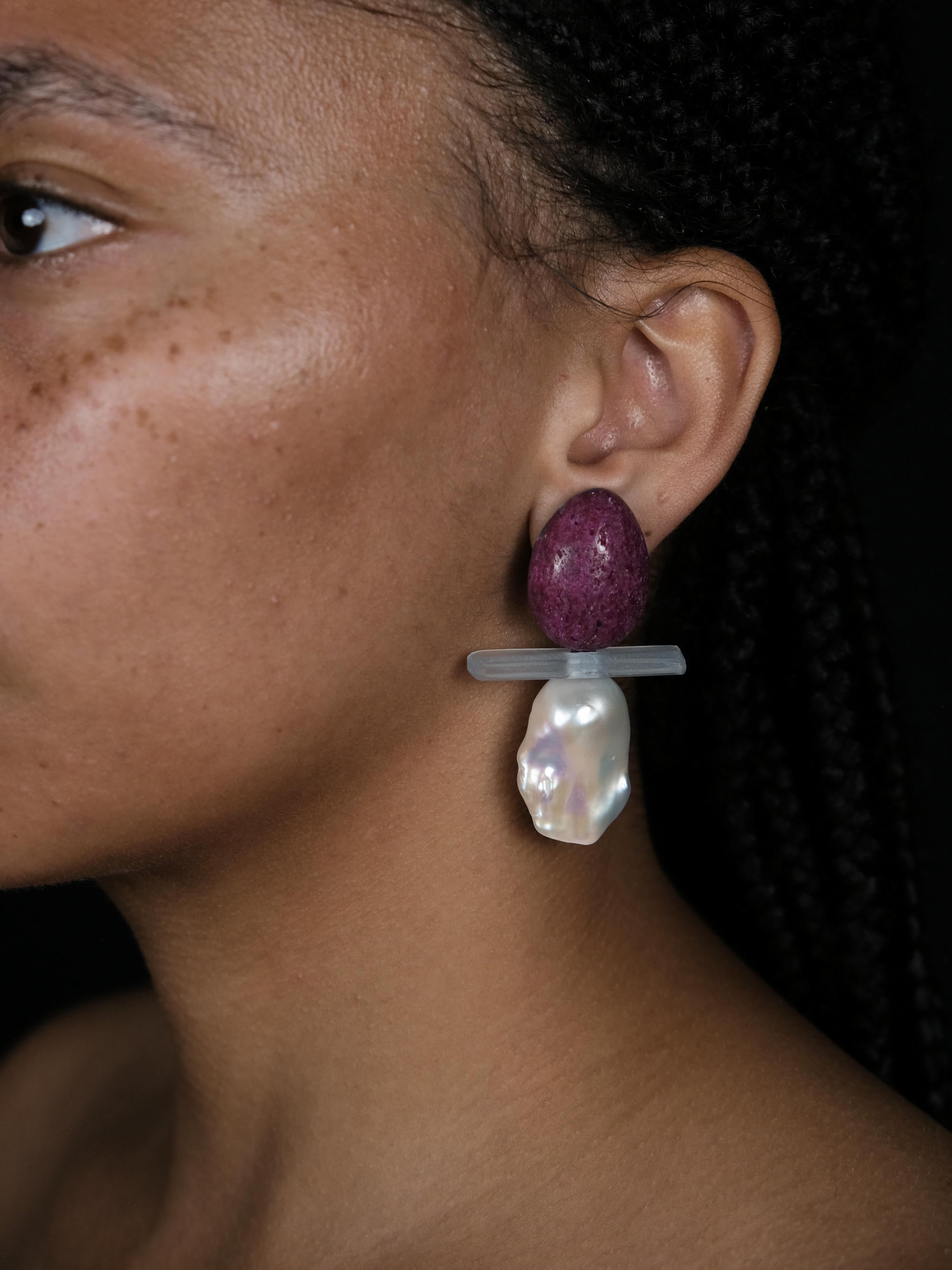A close-up of a person wearing a dangling earring with a textured purple top, a translucent gray horizontal bar, and a large white irregular pearl-like drop, against a dark background.