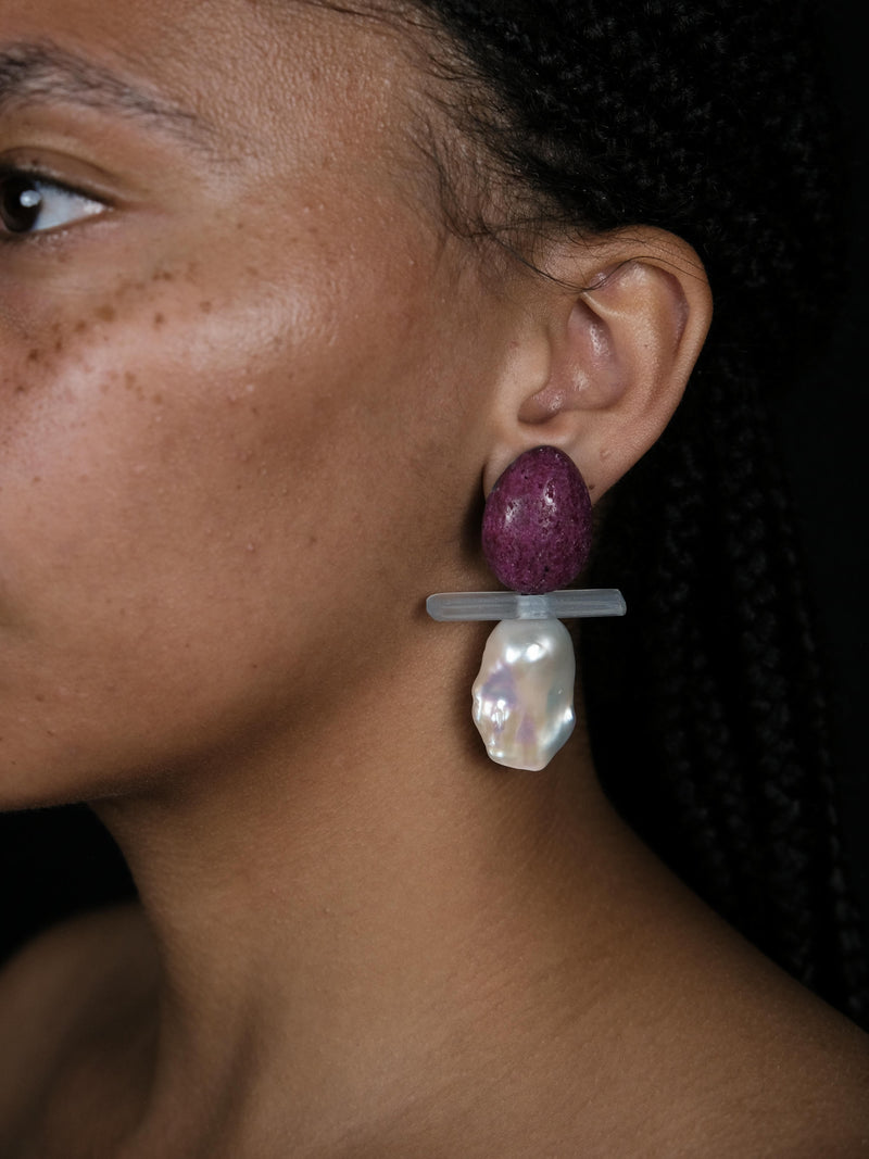 A close-up of a person wearing a dangling earring with a textured purple top, a translucent gray horizontal bar, and a large white irregular pearl-like drop, against a dark background.
