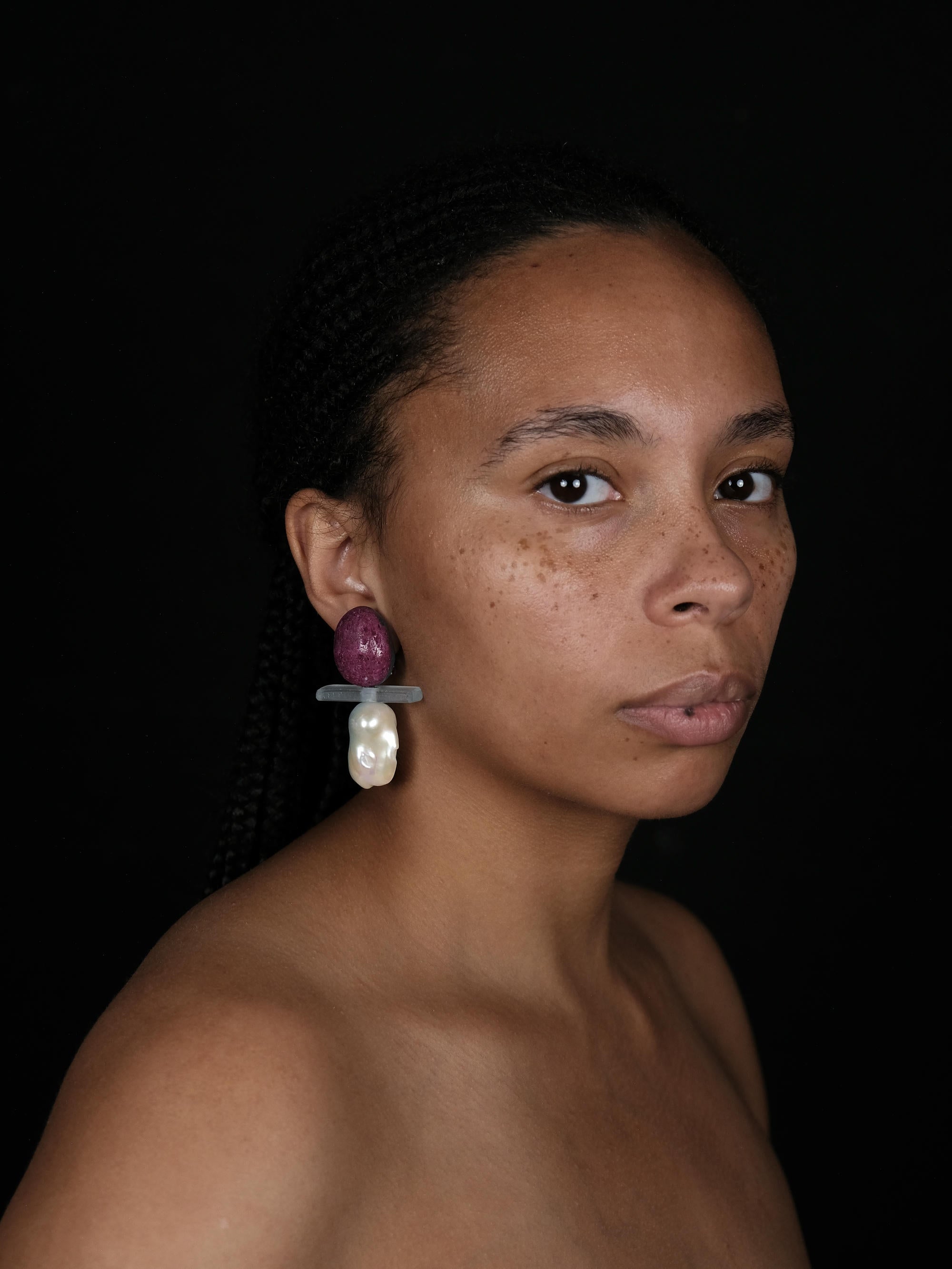 A close-up of a person wearing a dangling earring with a textured purple top, a translucent gray horizontal bar, and a large white irregular pearl-like drop, against a dark background.