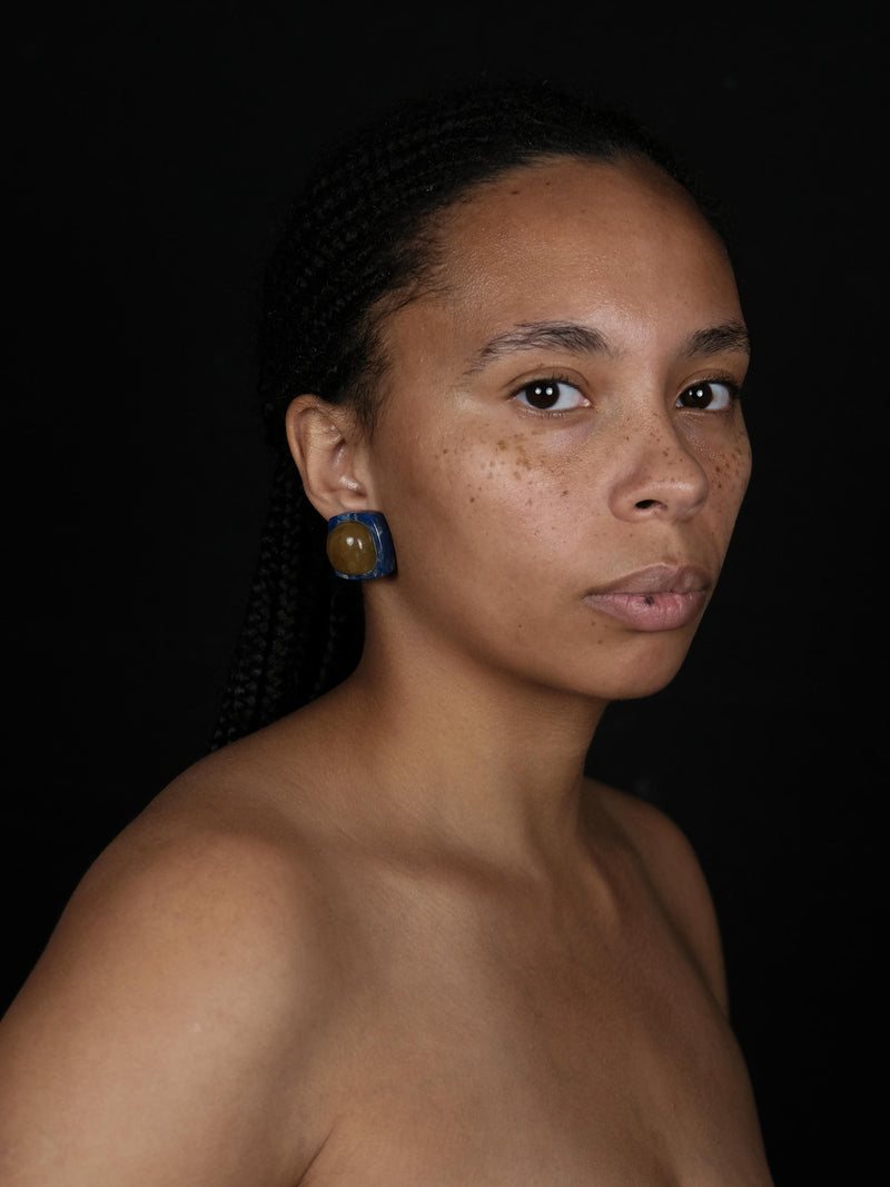 A woman wearing Earrings with rounded domeshaped yellow brownish stone set into a deep blue stone. Displayed on a white background. 