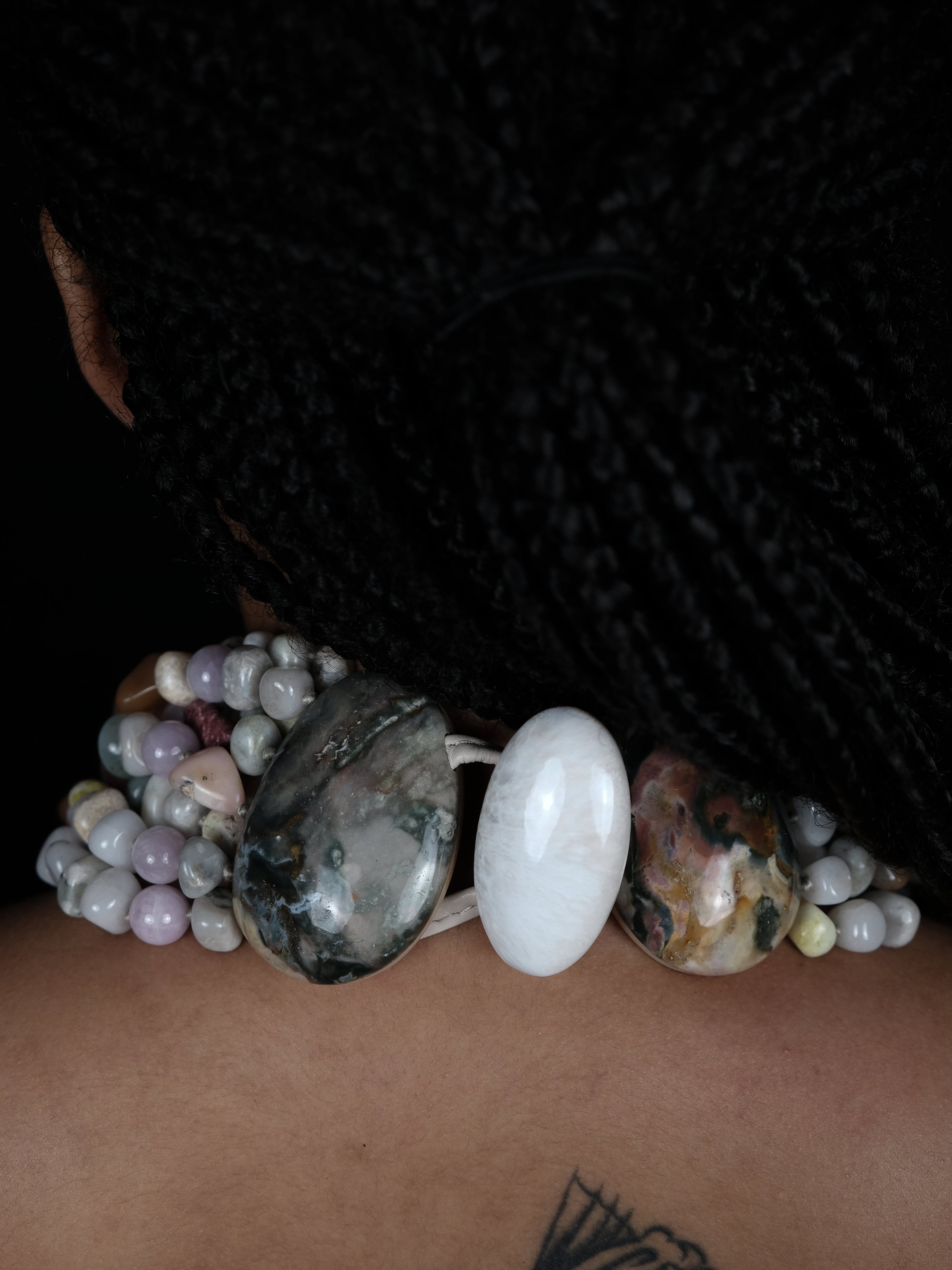 A close-up of a person's neck and upper chest wearing a multi-strand beaded bracelet with large polished stones, including a prominent white oval stone and a dark gray stone.