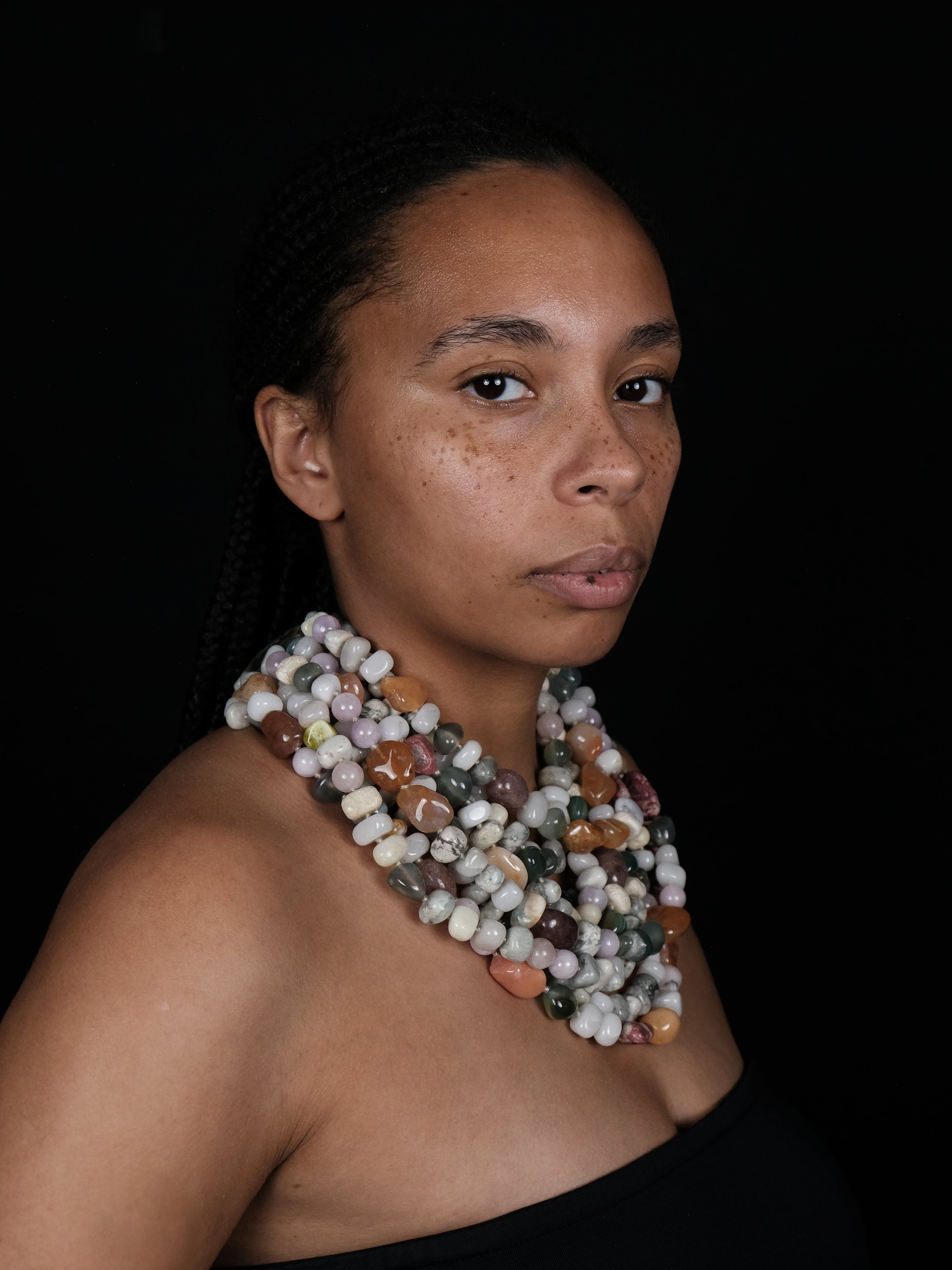 A woman with braided hair wearing a multi-strand statement necklace made of polished natural stone beads in earthy colors, set against a solid black background.