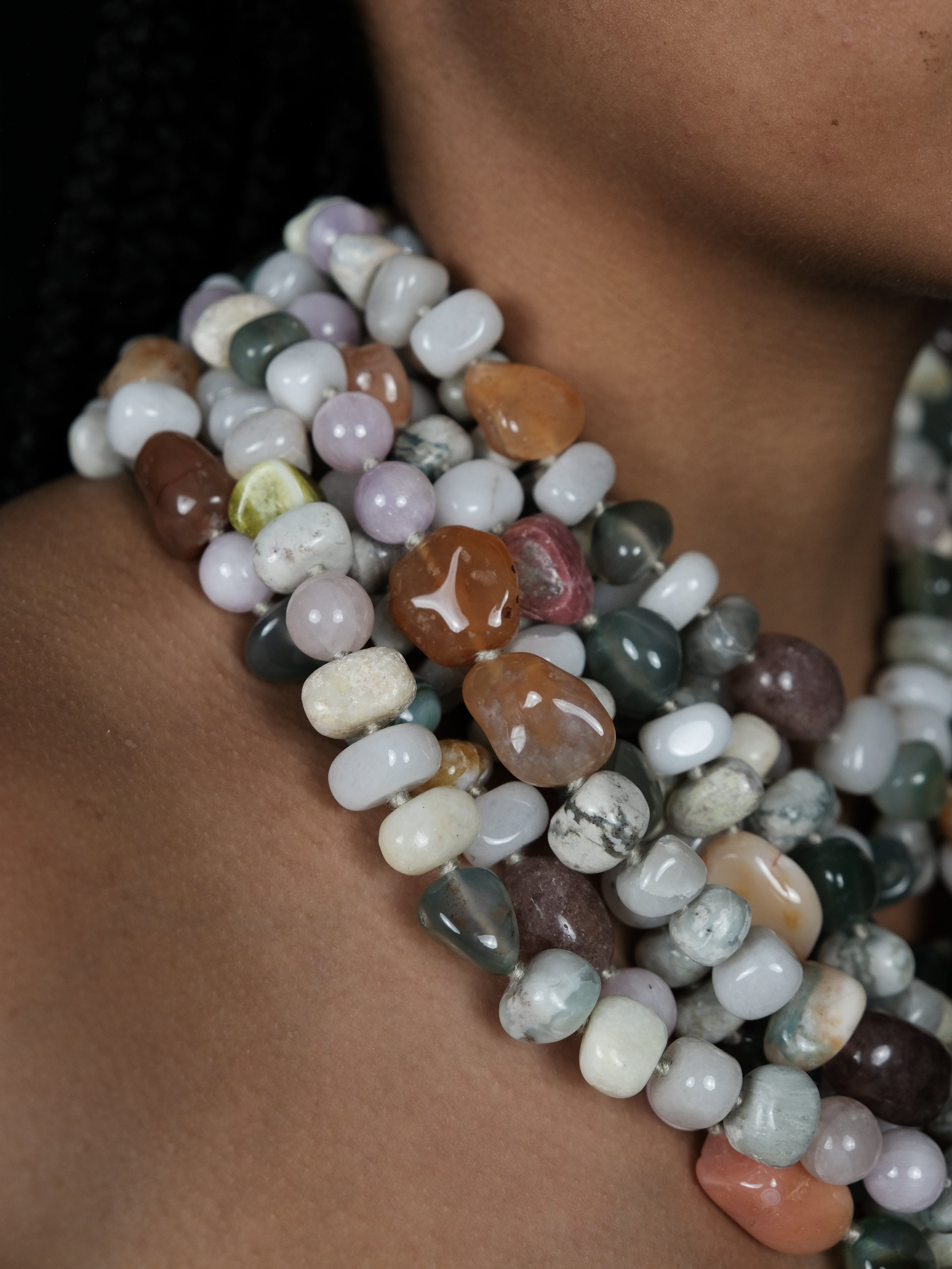 A close-up of a person wearing a multi-strand necklace made of polished, tumbled gemstone beads in various colors including white, gray, brown, green, and pink.