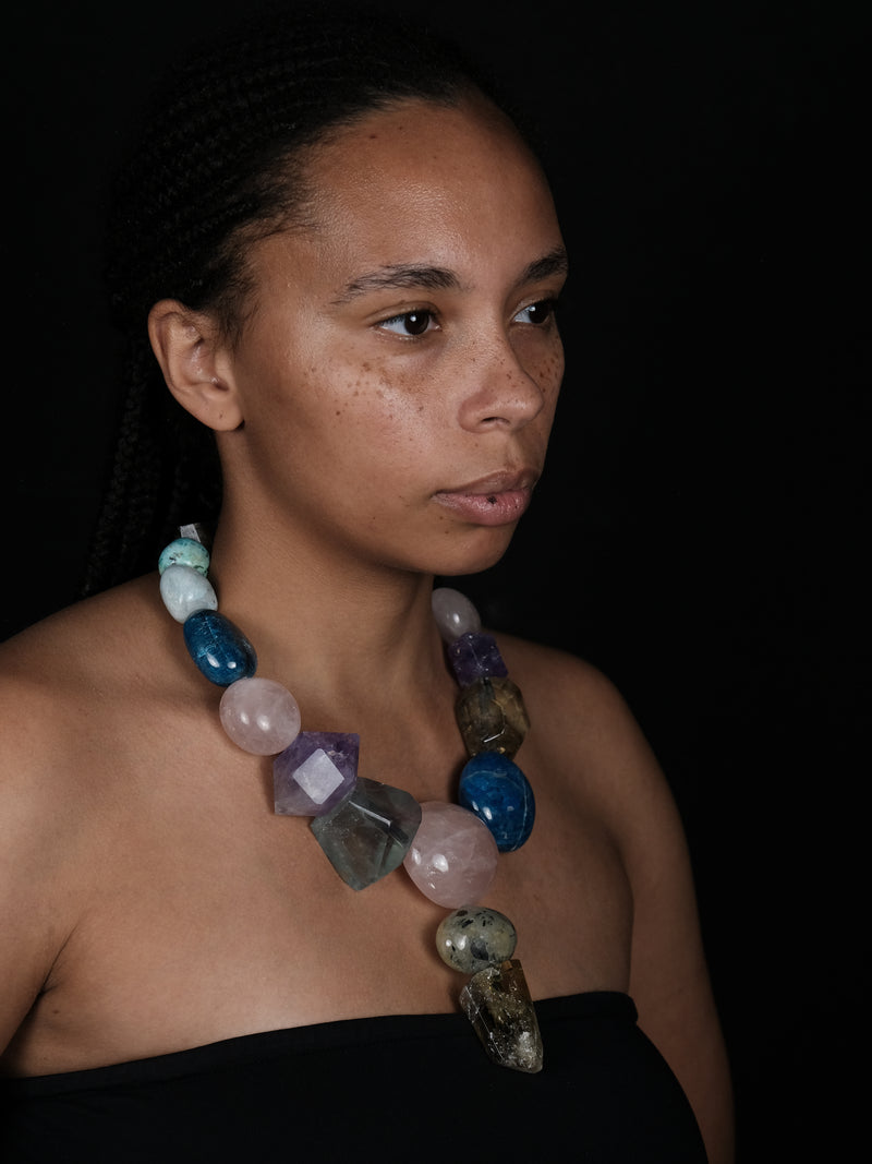 A woman wearing A multicolor beaded necklace featuring faceted gemstone beads in shades of pink, light blue, purple, amber, and clear, with a teardrop-shaped light blue stone pendant hanging from the center.