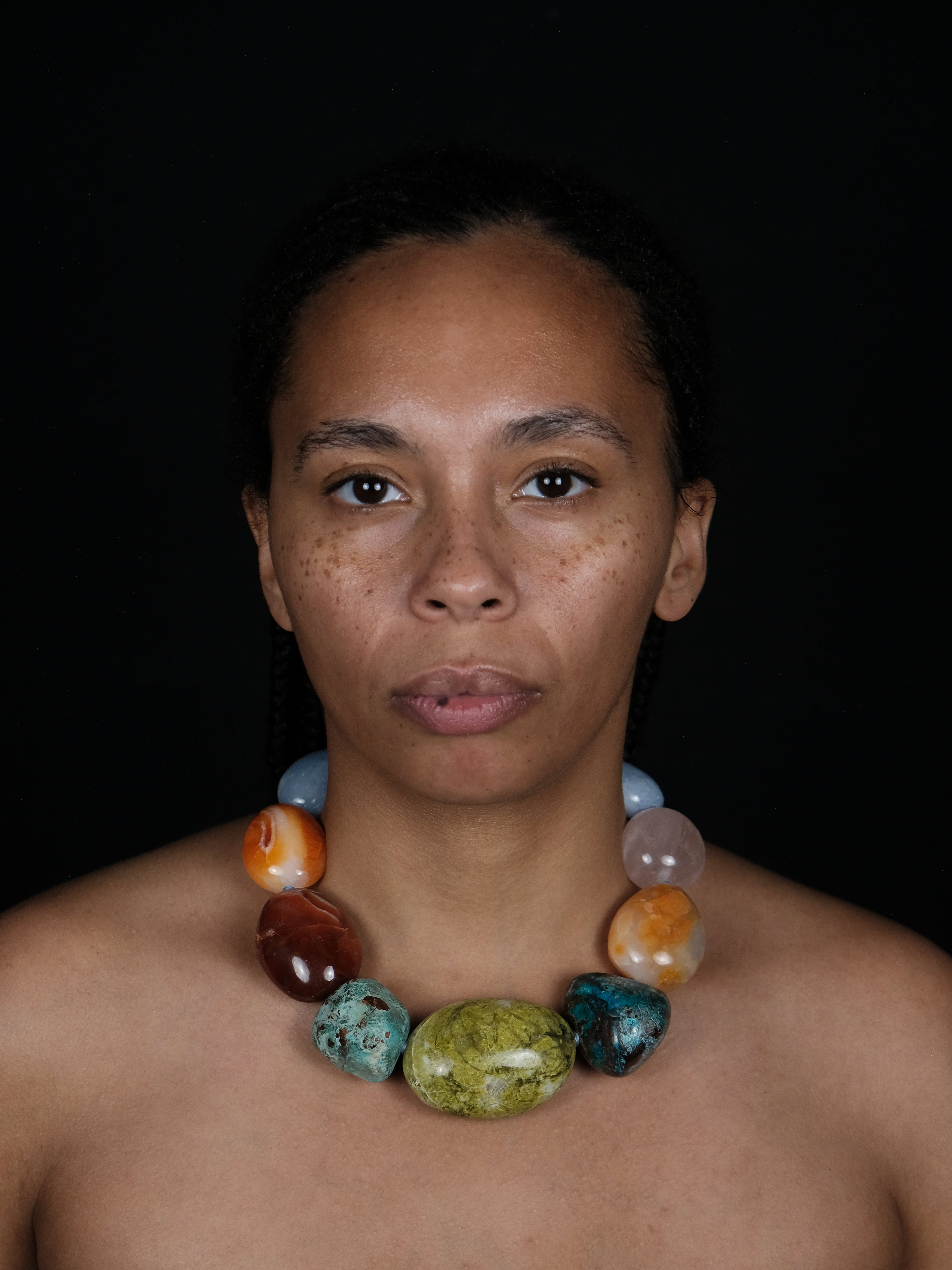 A close-up portrait of a woman wearing a chunky beaded necklace made of polished multicolored stones, set against a solid black background.