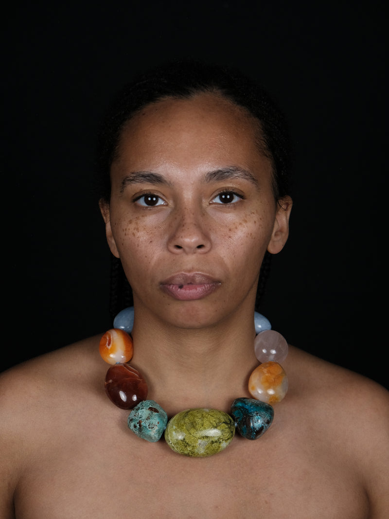 A close-up portrait of a woman wearing a chunky beaded necklace made of polished multicolored stones, set against a solid black background.