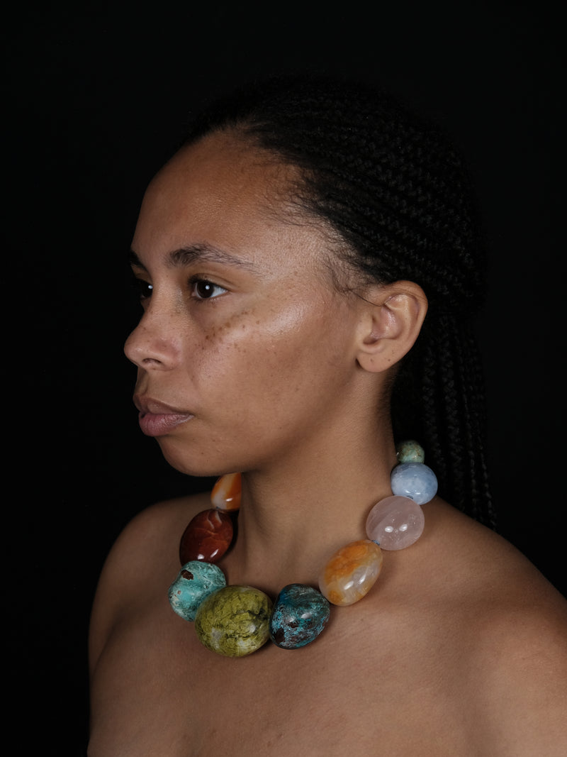 A close-up portrait of a woman wearing a chunky beaded necklace made of polished multicolored stones, set against a solid black background.