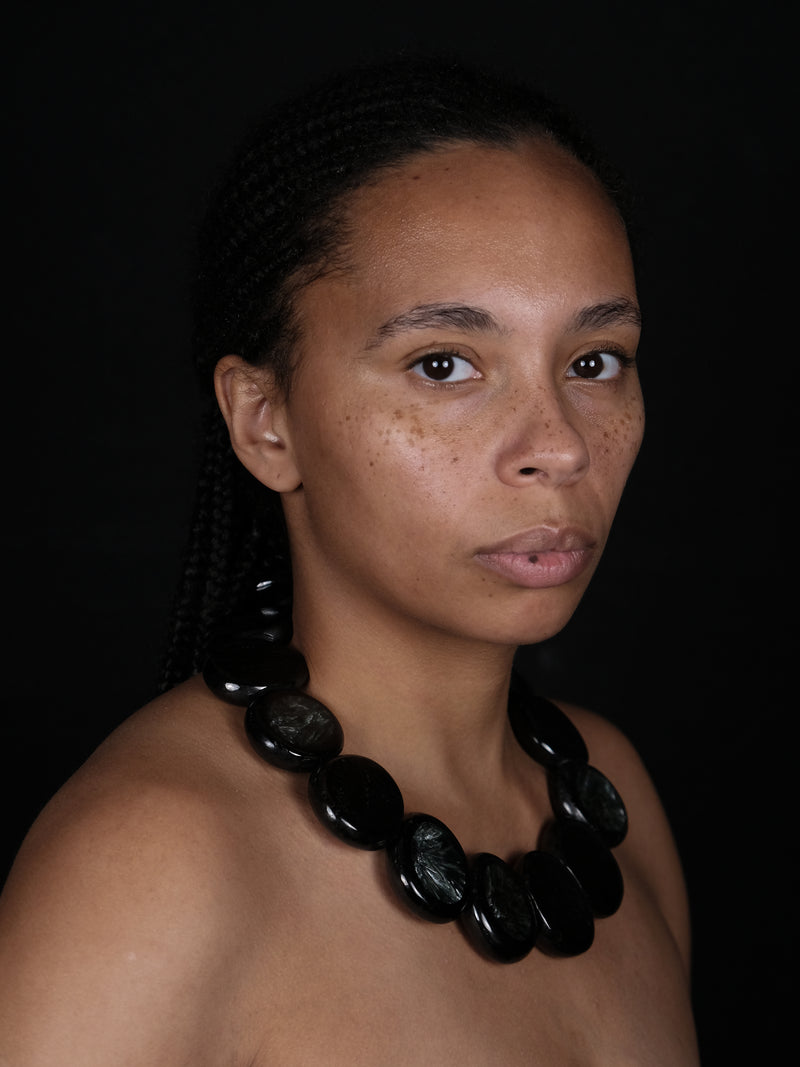 A close-up portrait of a woman with braided hair wearing a chunky black beaded necklace against a solid black background.