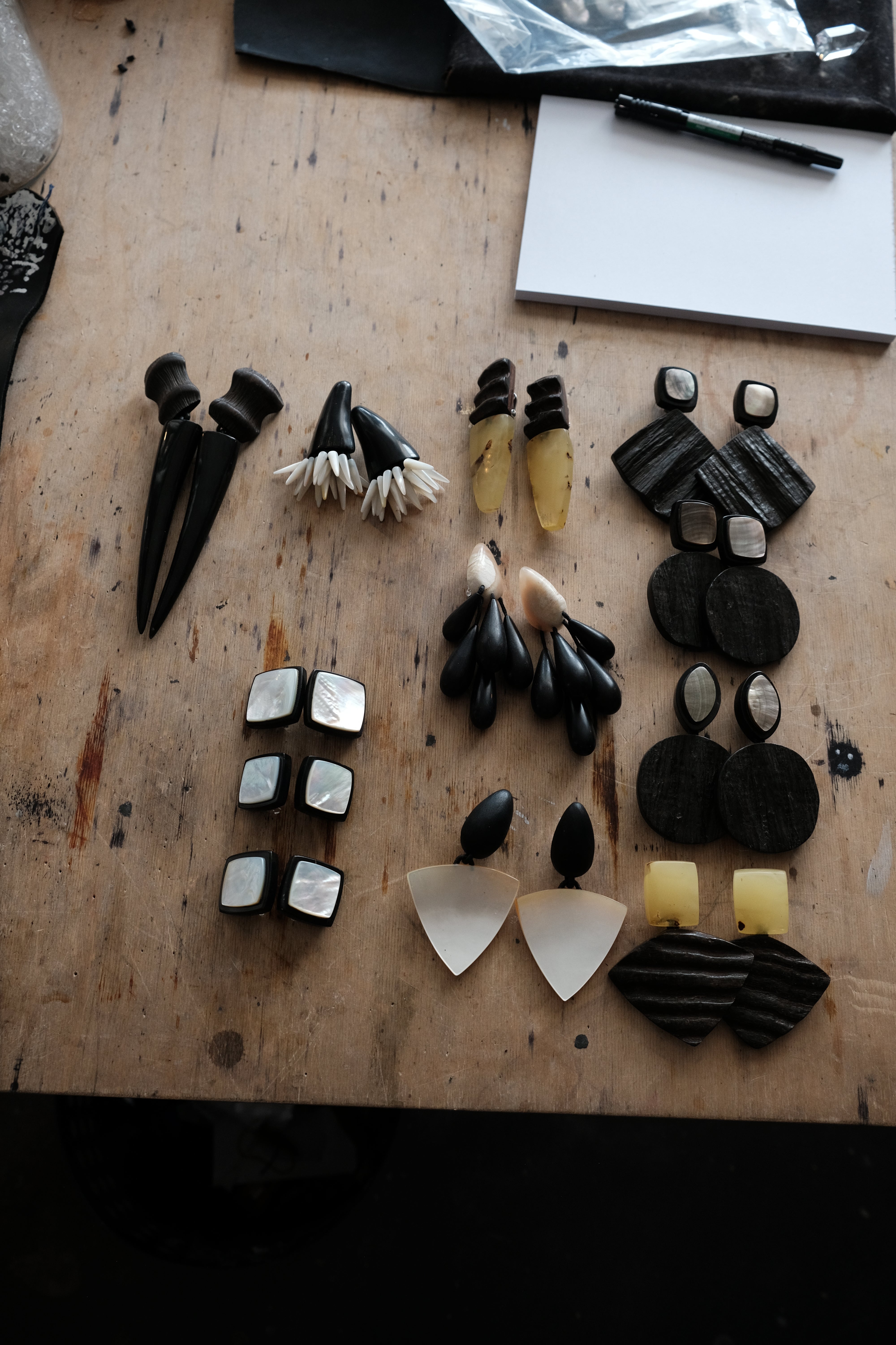 An assortment of handmade earrings and jewelry pieces arranged on a rustic wooden workbench. The collection features black, white, and natural materials including bone, shell, and wood.