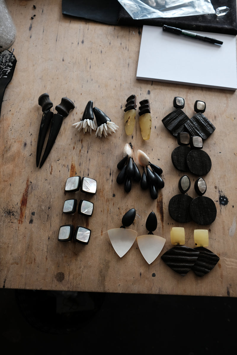 An assortment of handmade earrings and jewelry pieces arranged on a rustic wooden workbench. The collection features black, white, and natural materials including bone, shell, and wood.