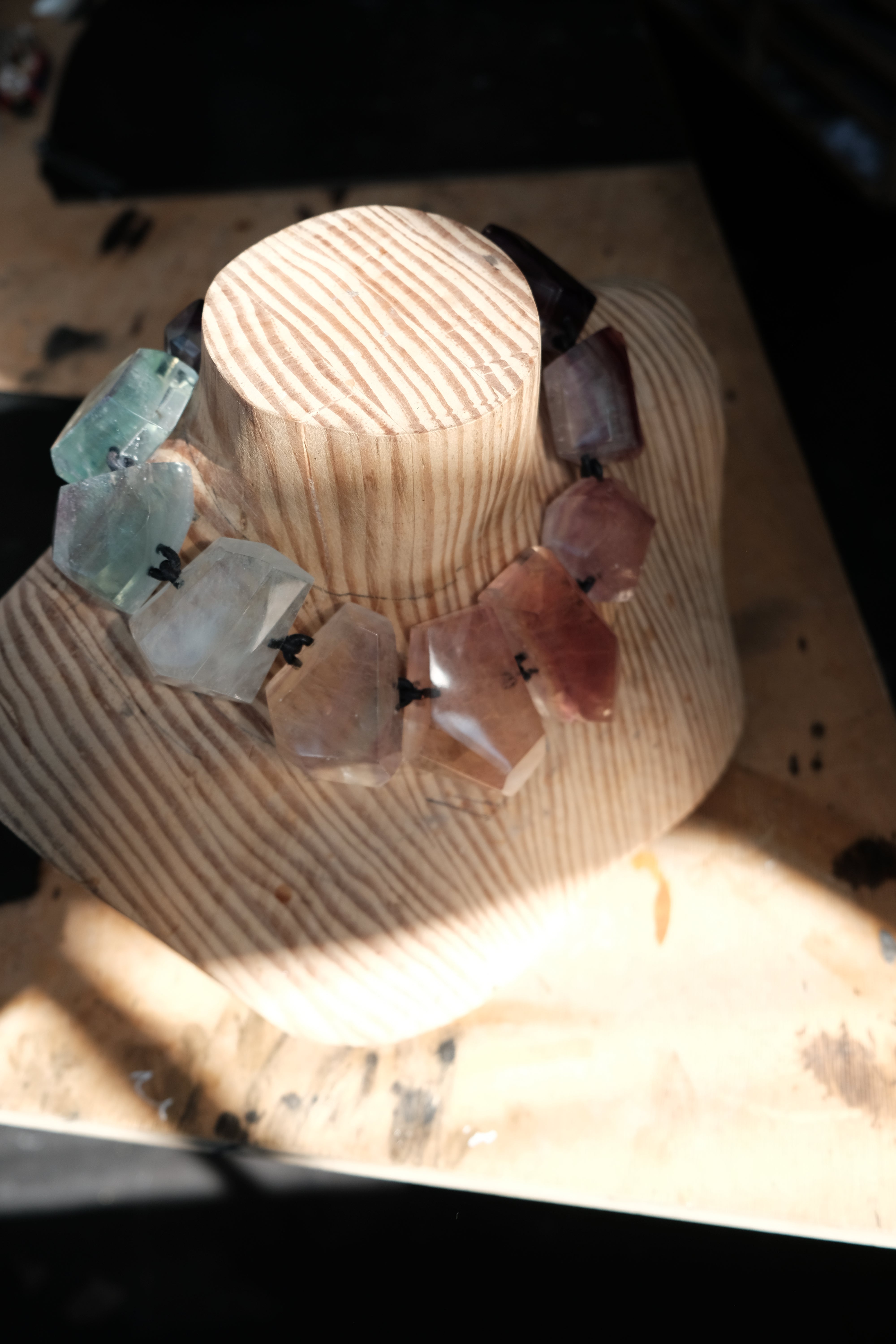 A handmade beaded necklace made of irregular, polished gemstone-like beads in shades of pink, purple, and light blue, displayed on a carved wooden stand.